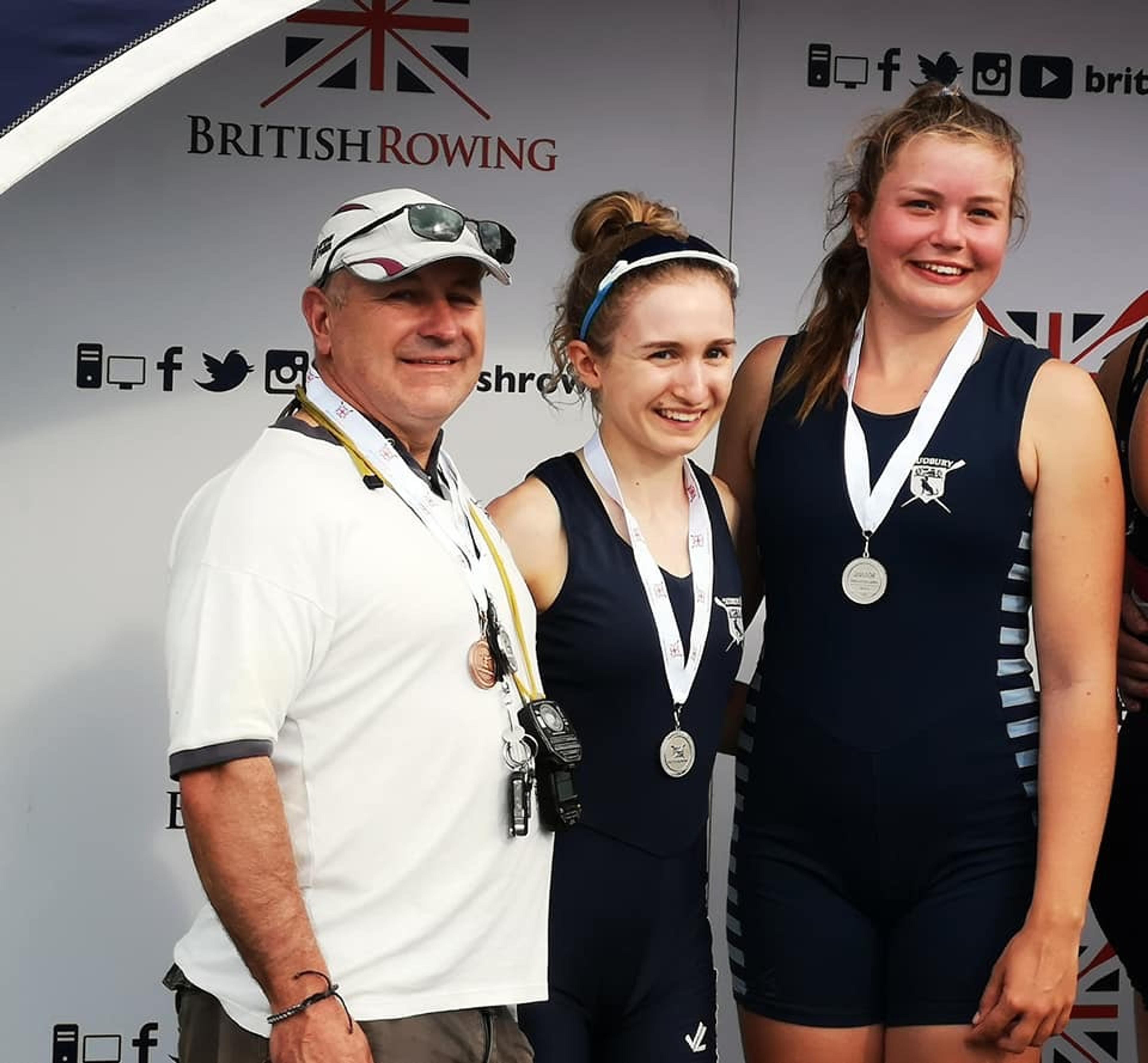 Wearing silver medals, the crew of women’s J18 double grins with their coach. 
