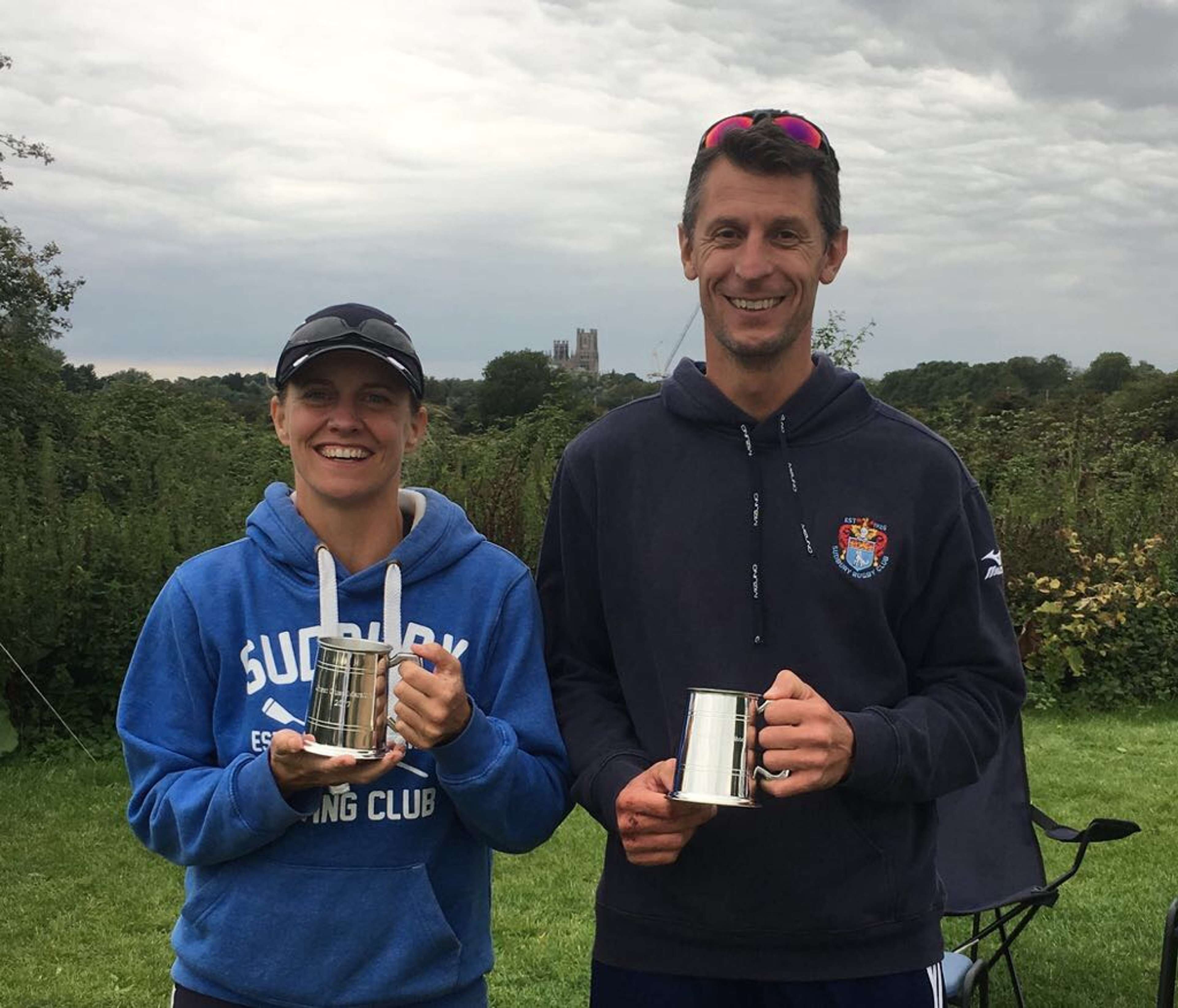 Two Sudbury rowers holding their pots (after winning a rowing marathon) with some sort of significant Lincolnshire ecclesiastical building in the distance in the far