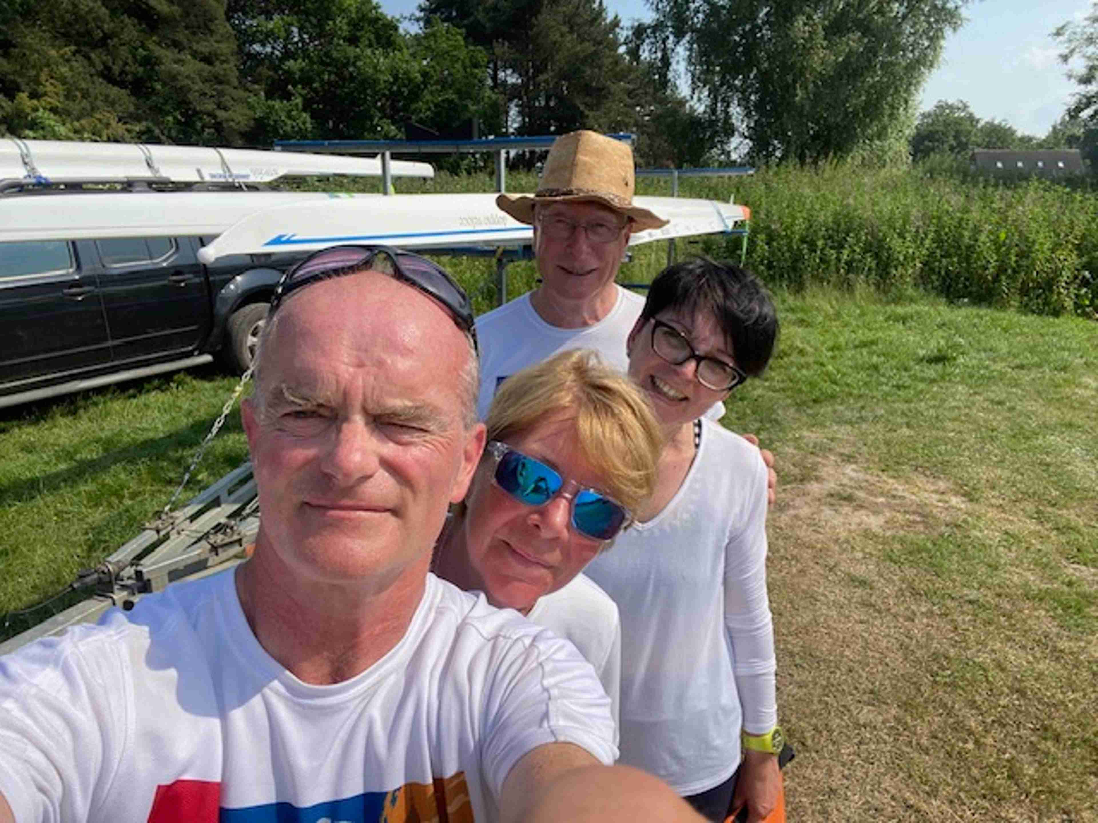 The crew take a selfie. Sean is in the foreground and taking the photo. Jeremy is in the distant background wearing a straw hat and reading glasses. Teresa and Tracy and queued up between them. Behind the four is a boat trailer, a bank of nettles and a line of trees.