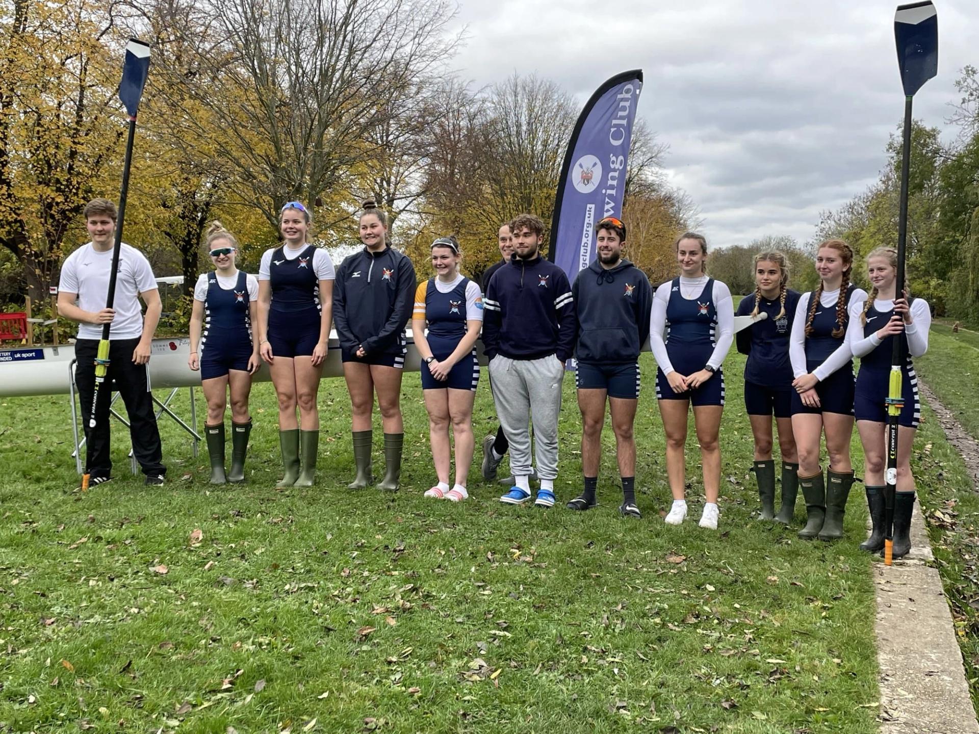 The twelve Sudbury rowers pose for a photo at their grassy boating point. It looks chilly and autumnal.