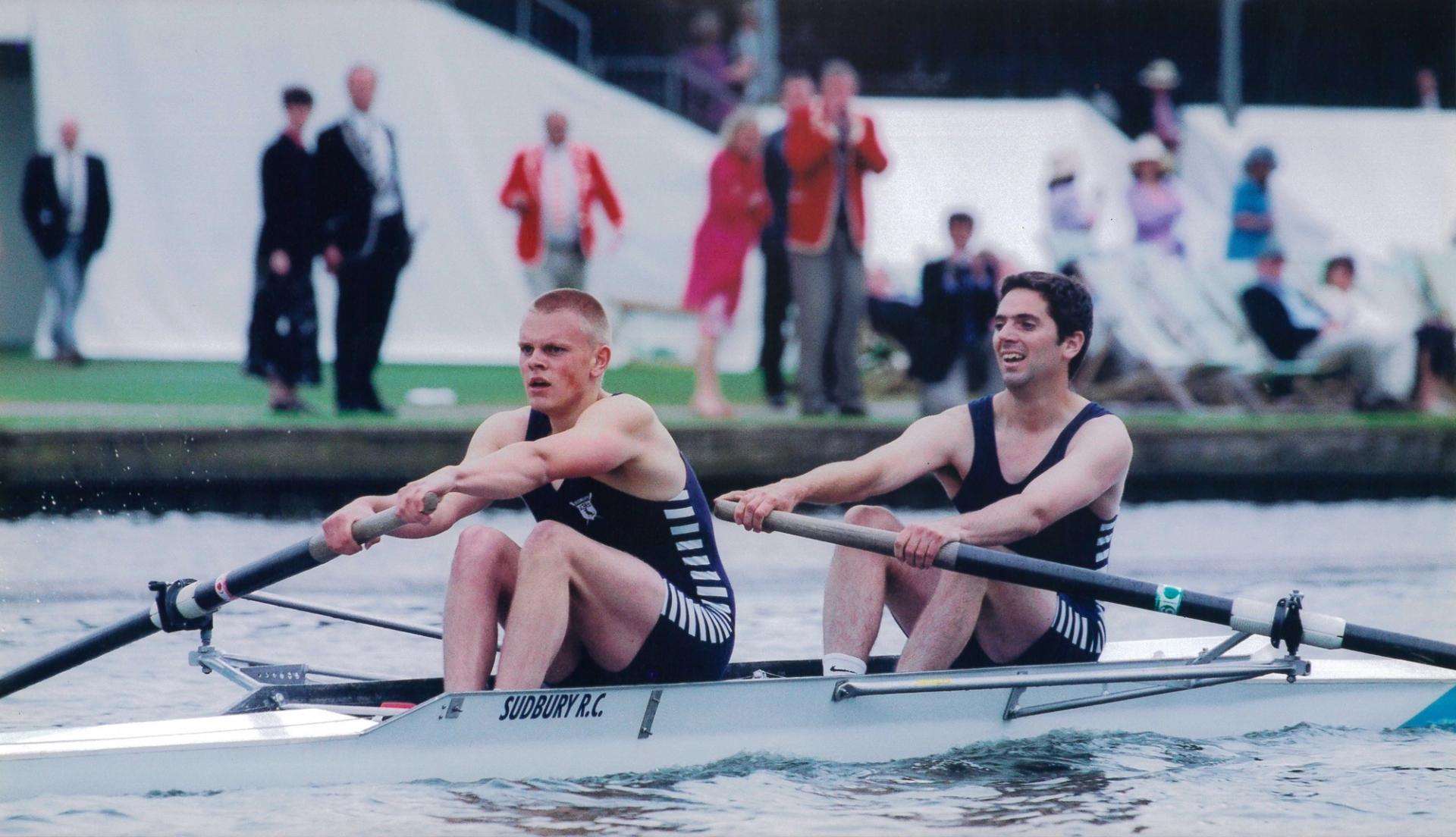 Two male rowers from Sudbury Rowing Club competing in a pairs event. They are mid-stroke in a white racing shell, wearing dark uniforms with white stripes. The rower in front has short blond hair and looks intensely focused. Behind them, spectators in colorful attire watch from the riverbank. The scene suggests a formal rowing regatta, possibly at Henley.