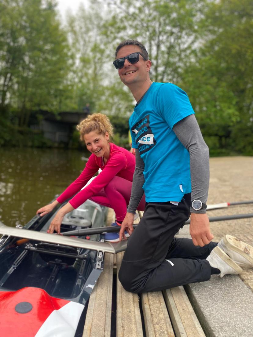 A photo taken at ground level on the club’s wooden landing stage. A training scull is in the water, help by a smiling volunteer as a beginner tentatively gets into the boat. 