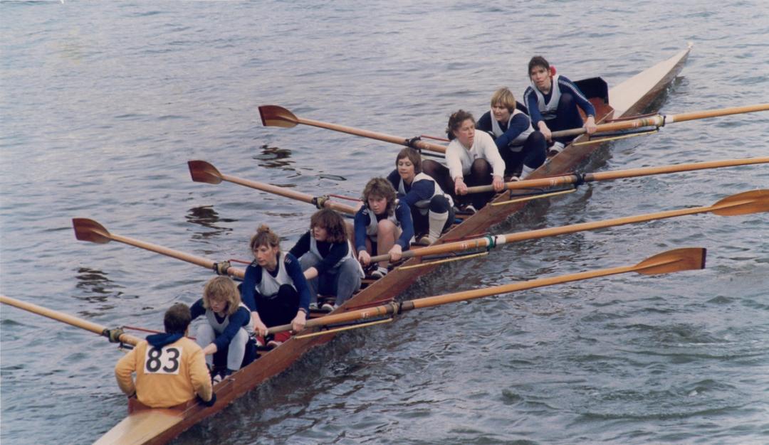  A rowing crew mid-race on a river. The crew consists of eight women, each with an oar, and a cox at the stern wearing a yellow jacket with the number 83. The rowers wear matching blue and white athletic uniforms. The boat cuts through the water, with the rowers synchronised in their efforts. The scene appears lively and focused.