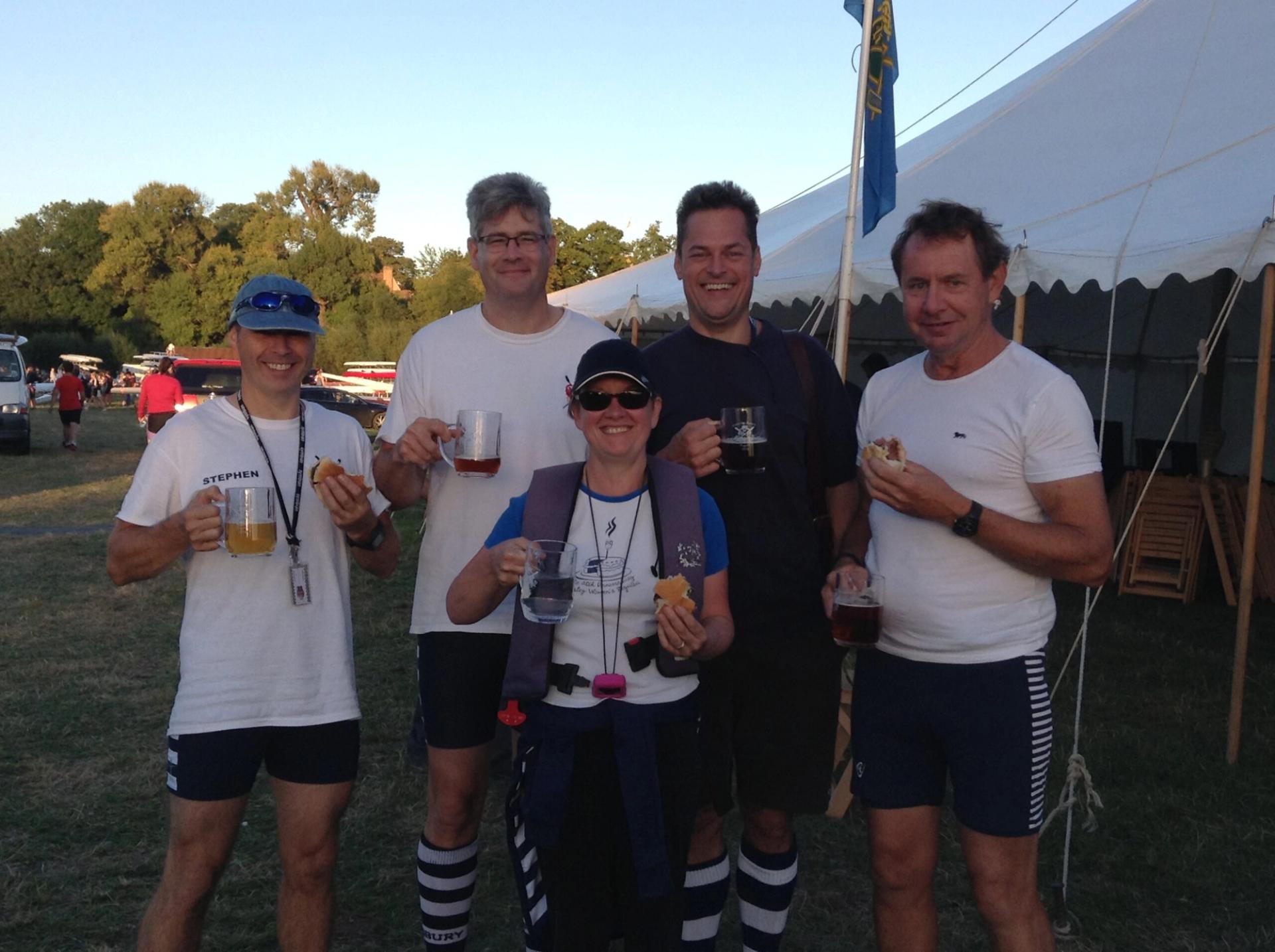 Four rowers and their cox, all holding nearly-empty beer tankards and posing outside a rustic marquee.