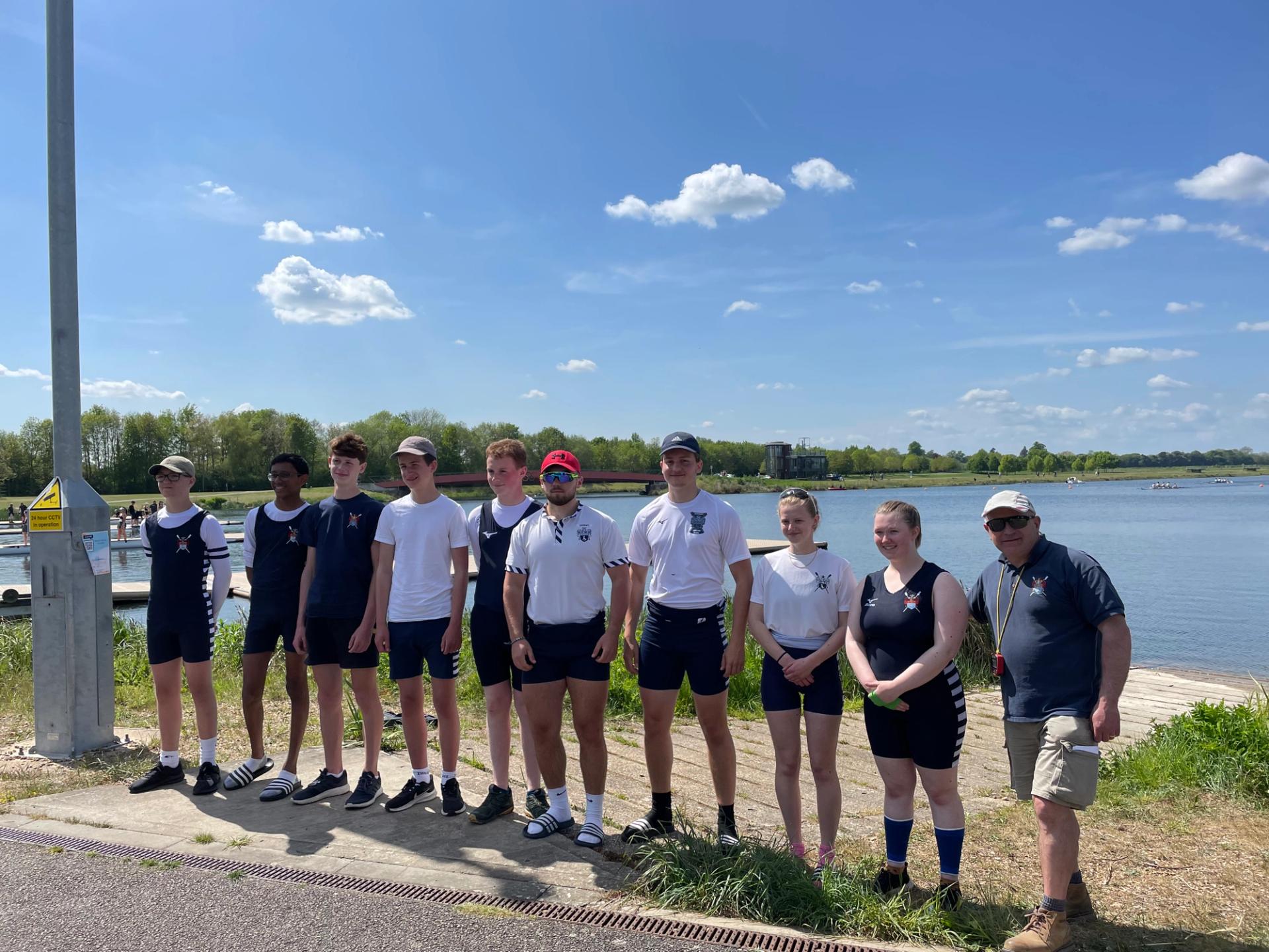 Nine juniors and a coach standing in the sunshine in front of the vast rowing lake at Dorney. 