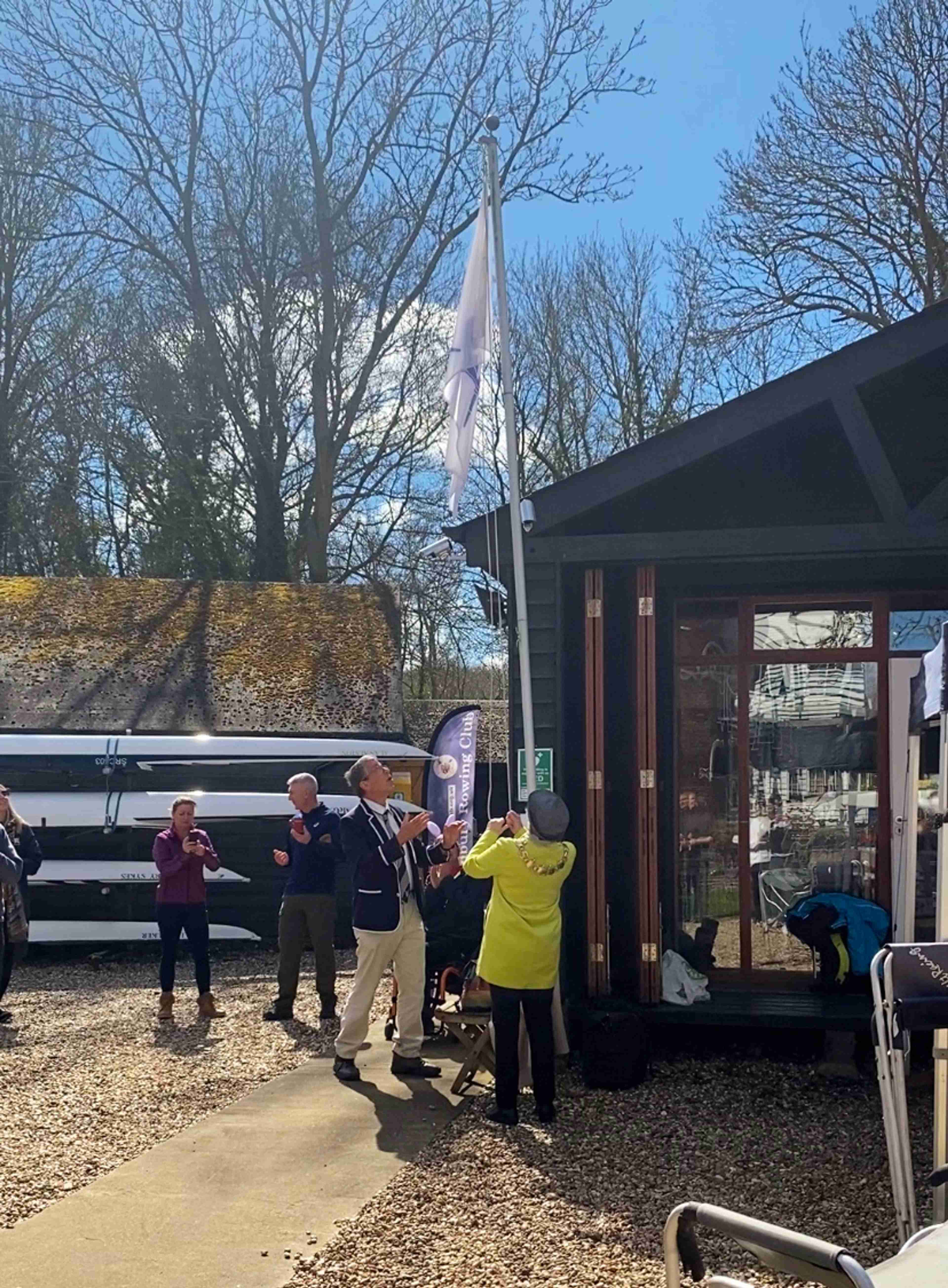 A small group of people standing on the gravel outside the clubhouse, watching as the lady mayor (who wears a distinctive, long yellow coat) raises the club flag up the pole. Andrew is standing very close to the pole, looking at the flag, palms outsretched and mouth agog in genuine wonder.