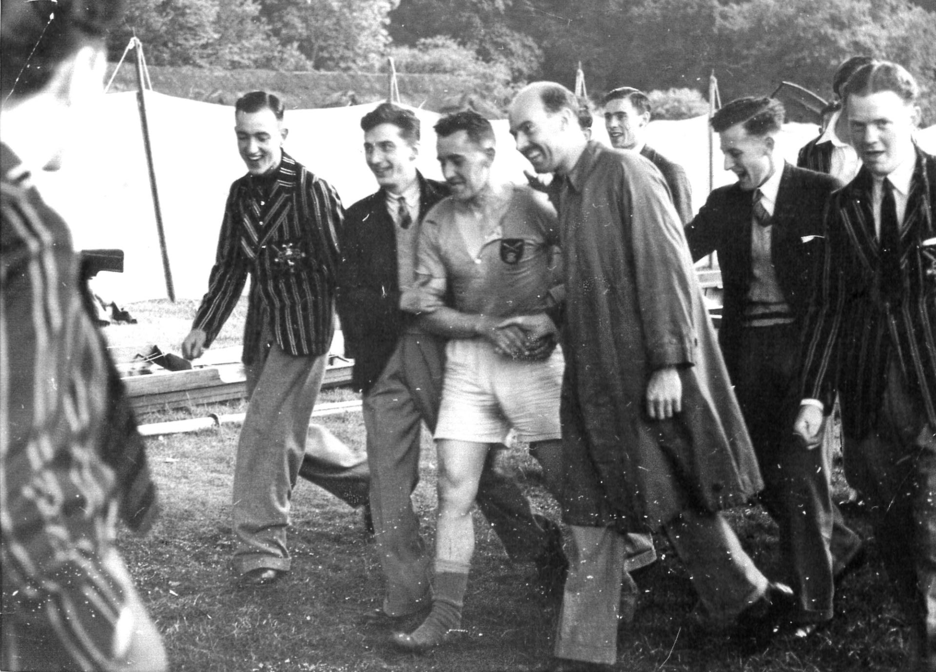 A black and white photograph from the mid-20th century showing a group of young men, likely a rowing team, standing together outdoors. One man wears a striped blazer, another is in shorts and athletic wear, while others are in suits or casual jackets. They appear to be at a sporting event, with tents visible in the background. The men are smiling and seem to be in good spirits, suggesting a camaraderie typical of team sports.