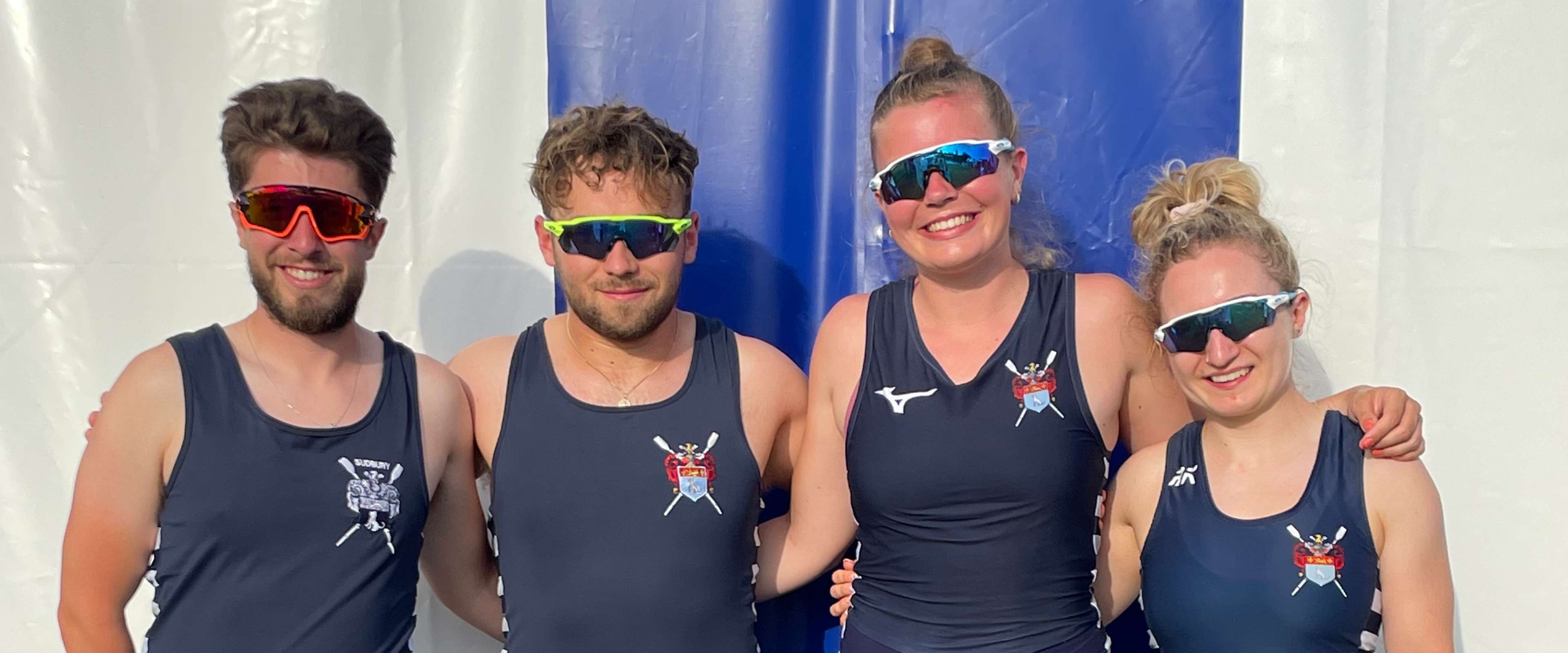 Byron Bullen, Harry Moule, Martha Bullen and Amelia Moule pose for a photo, arms linked, wearing sunglasses and all-in-ones in front of a tent at Henley.