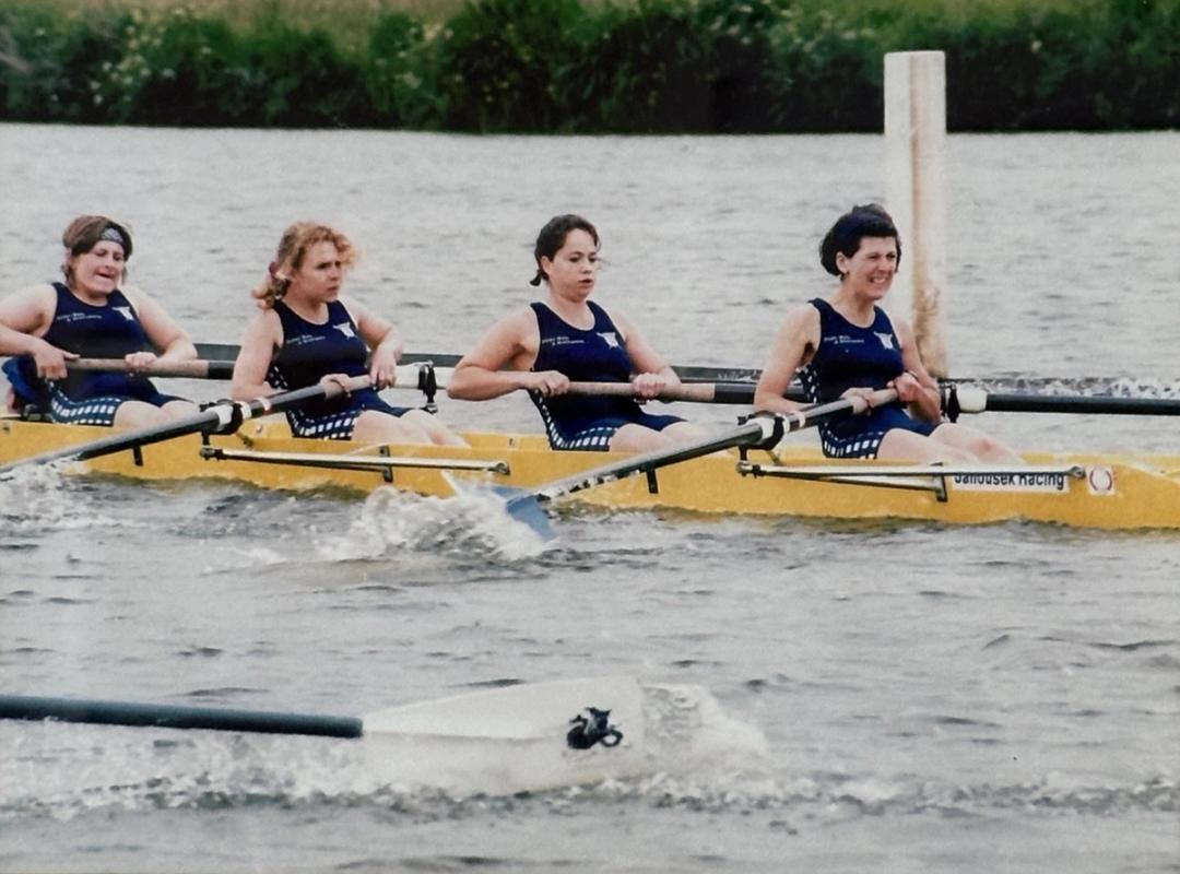 A women's rowing crew competing in the 1995 Henley Women's Regatta. Four women are seen rowing a yellow boat on a river, wearing matching dark blue uniforms. 