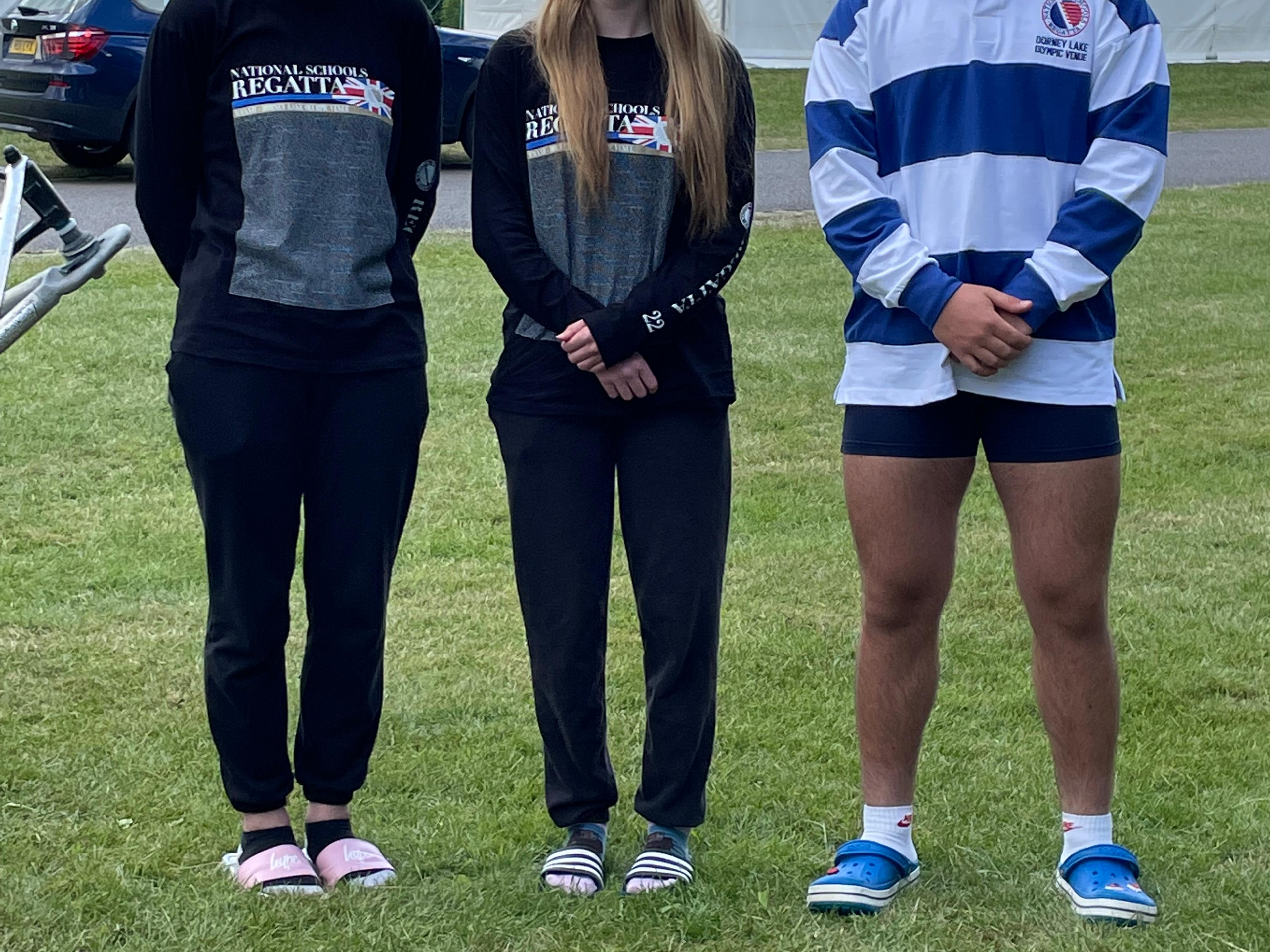 Three junior rowers standing on the grass outside a pair of white tents at a rowing event.