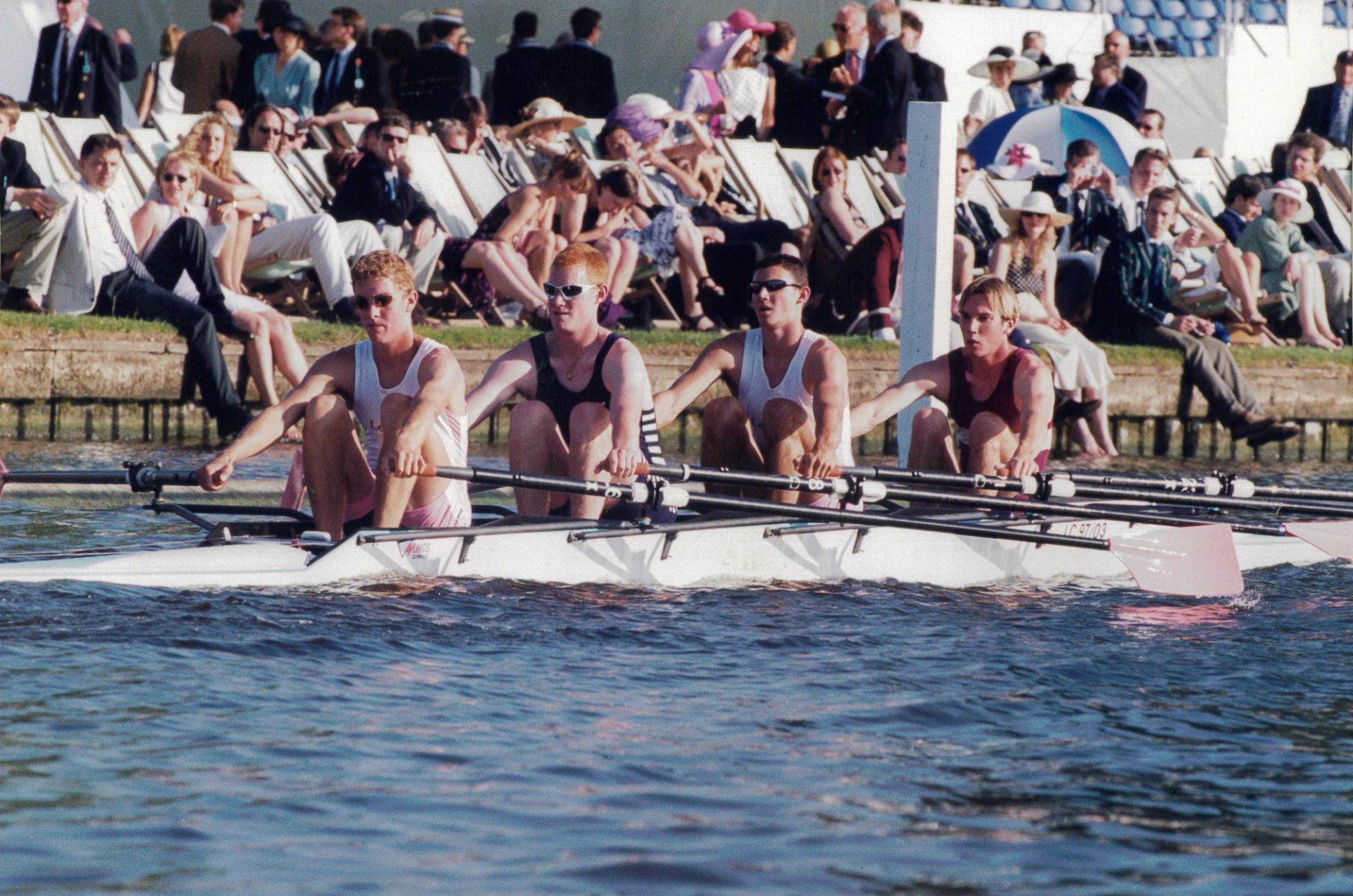 A rowing crew of four young men competing in a regatta. They are seated in a white racing shell on the water, wearing alternating white and dark tank tops. Behind them, a crowd of spectators sits in deck chairs along the riverbank, many wearing formal attire and hats. The scene suggests a prestigious rowing event like Henley Royal Regatta.