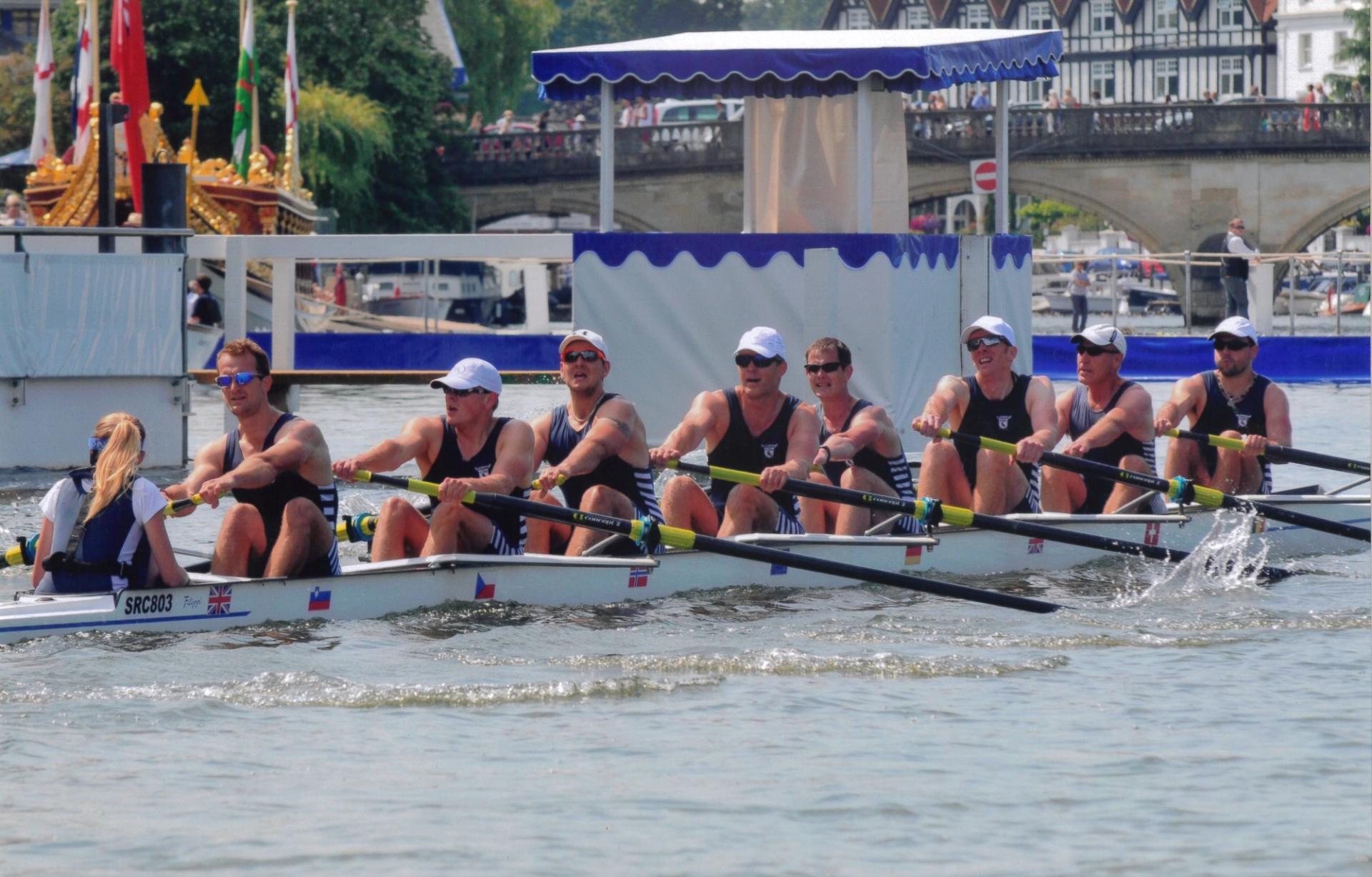 A men's eight rowing crew competing at what appears to be the Henley Royal Regatta. The rowers, wearing dark uniforms with white stripes, are mid-stroke in a white racing shell marked with various national flags. A coxswain is visible at the front. In the background, there's a bridge with spectators, traditional Tudor-style buildings, and part of an ornate boat, likely the umpire's launch. A blue and white canopied structure is visible on the riverbank, typical of the Henley Regatta enclosures.