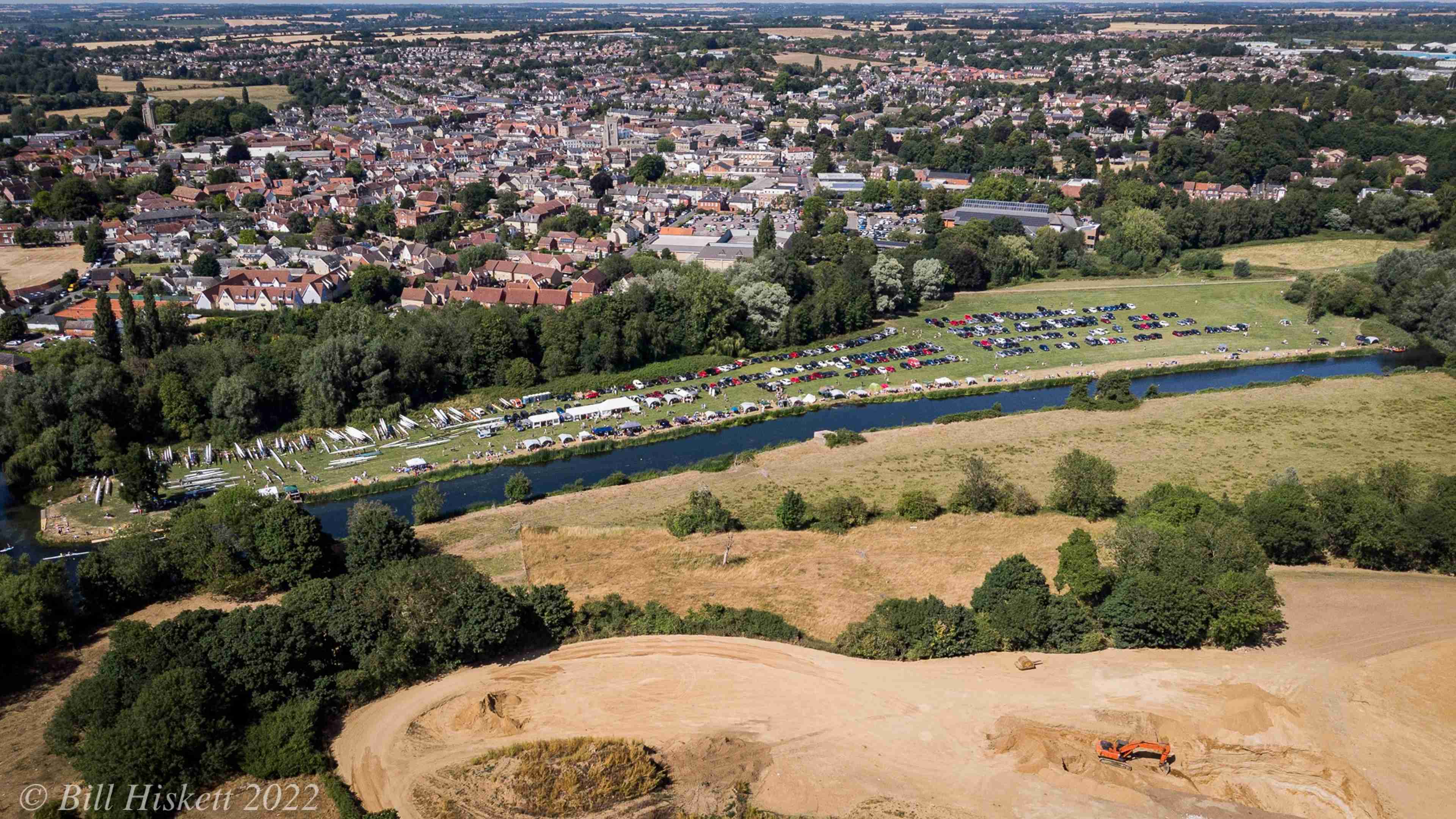 An aerial shot of the meadow, where there are boats, cars, white tents and spectators, with Sudbury town in the background.