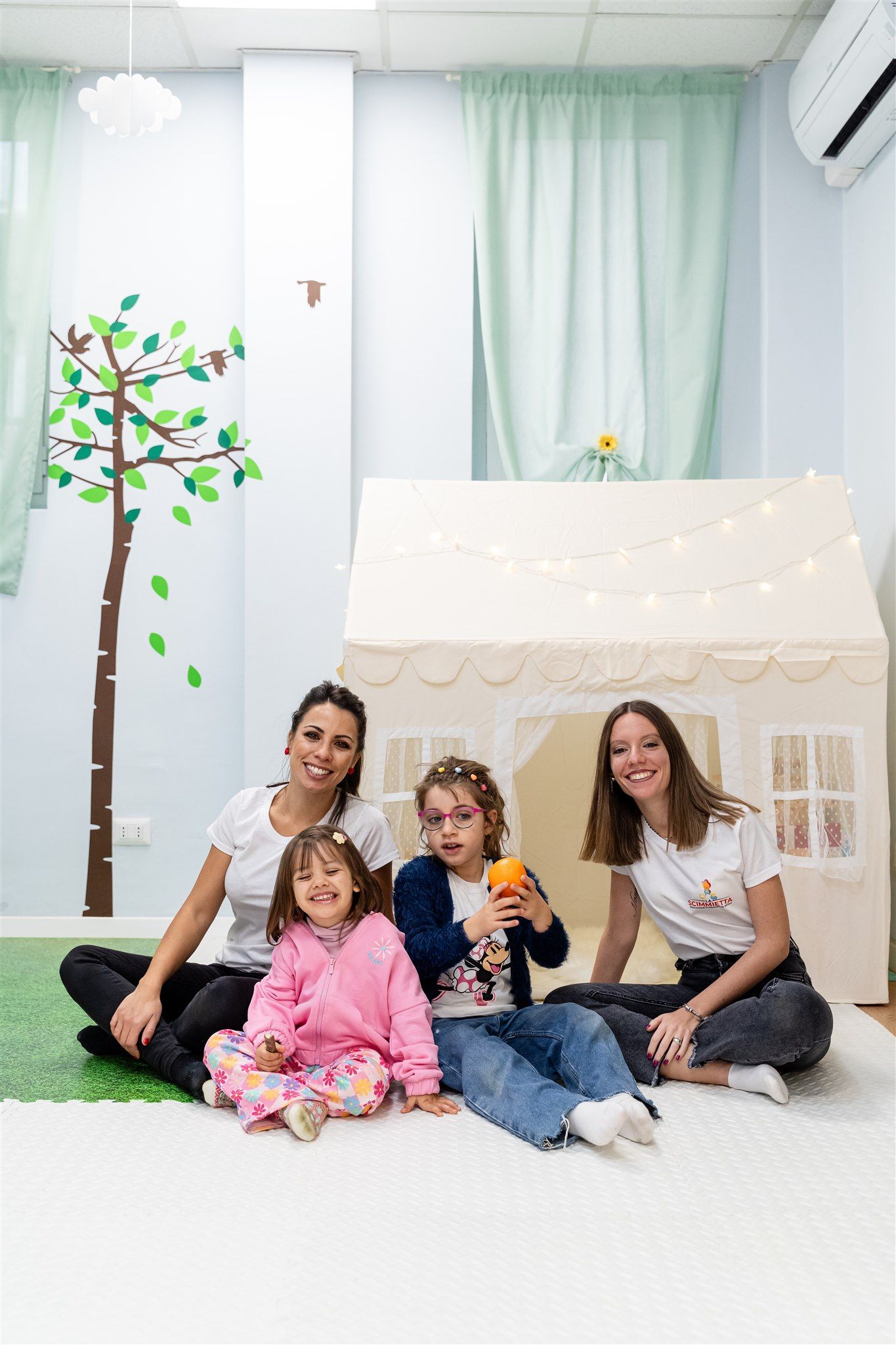 Due educatrici della Scimmietta sedute sul tappeto con tre bambini, davanti alla casetta di stoffa della ludoteca