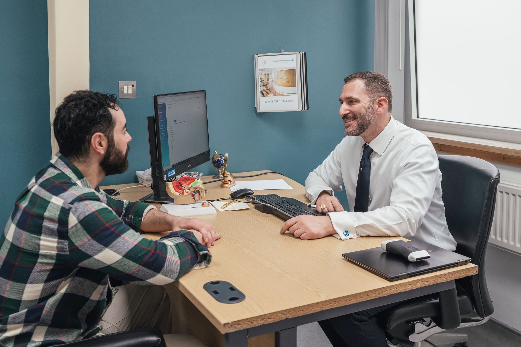 Mr Ollandini and a patient sit facing each other across a desk during a consultation in a medical clinic. Mr Ollandini, dressed in a white shirt and tie, is smiling warmly and speaking with the patient, who is wearing a green and navy checked shirt. A computer screen, anatomical prostate model, and small desk items are visible on the table, creating a professional and welcoming clinical environment.