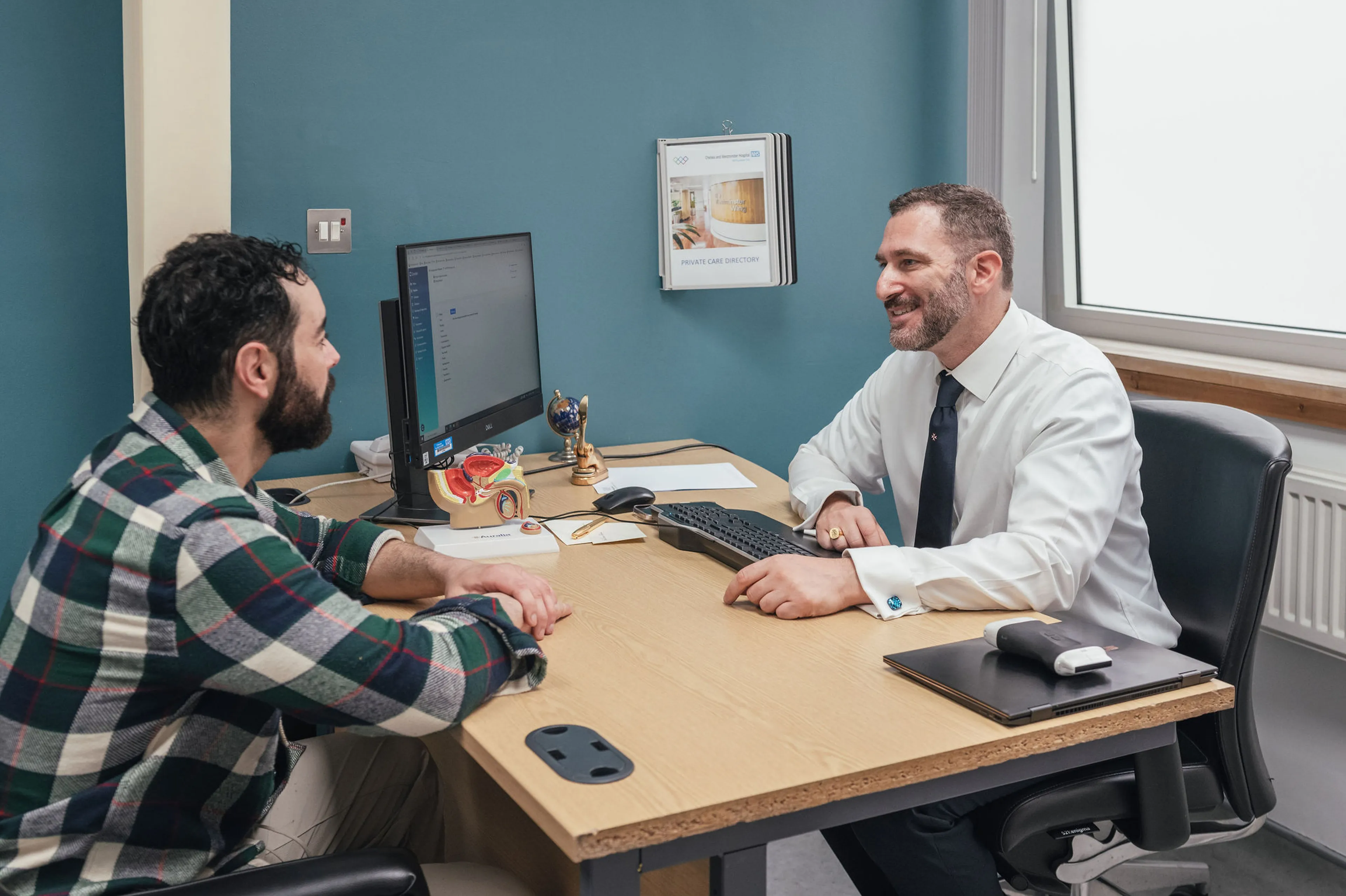 Mr Ollandini and a patient sit facing each other across a desk during a consultation in a medical clinic. Mr Ollandini, dressed in a white shirt and tie, is smiling warmly and speaking with the patient, who is wearing a green and navy checked shirt. A computer screen, anatomical prostate model, and small desk items are visible on the table, creating a professional and welcoming clinical environment.