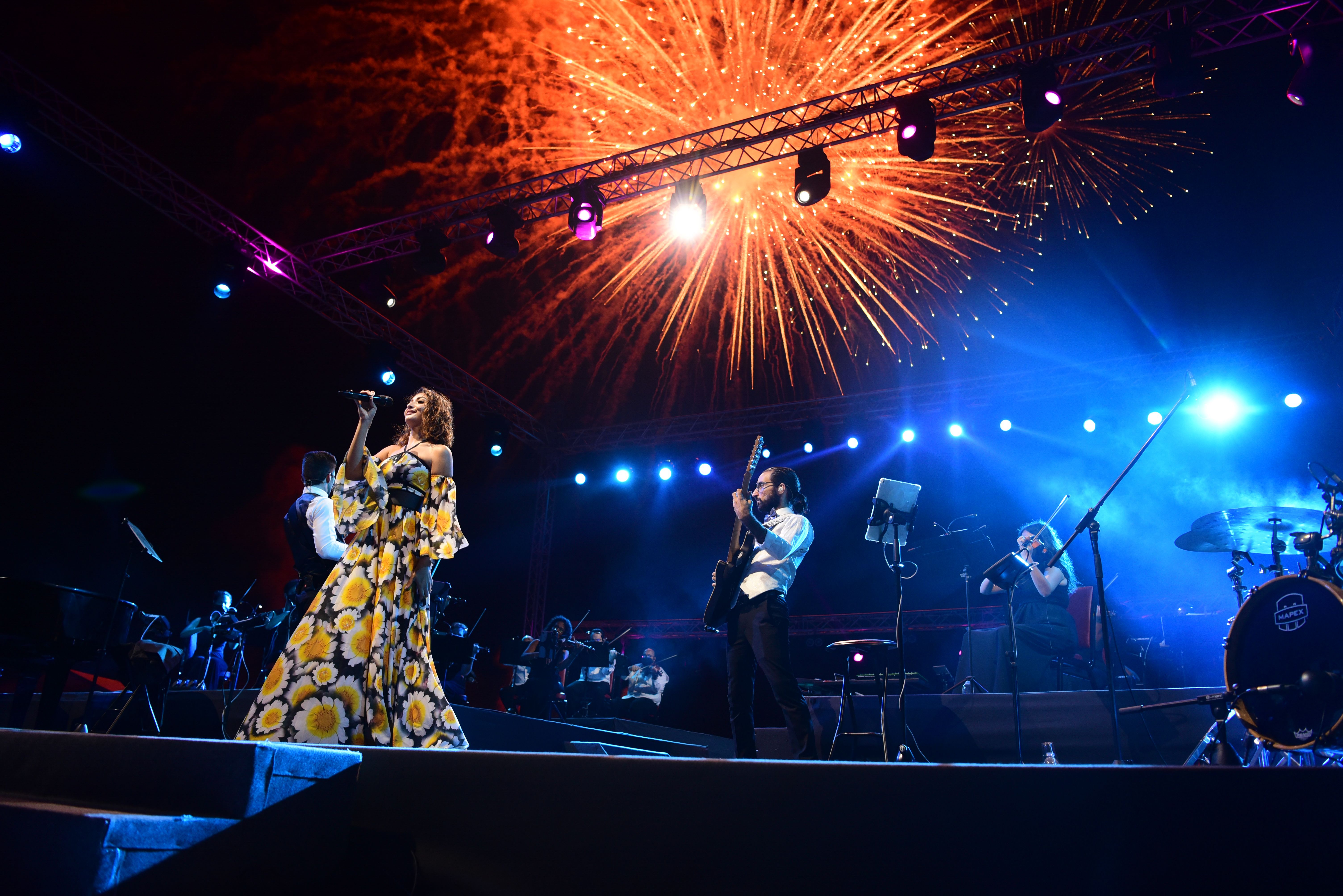 a woman in a floral gown sings while a guitarist plays behind her, under a bursting firework