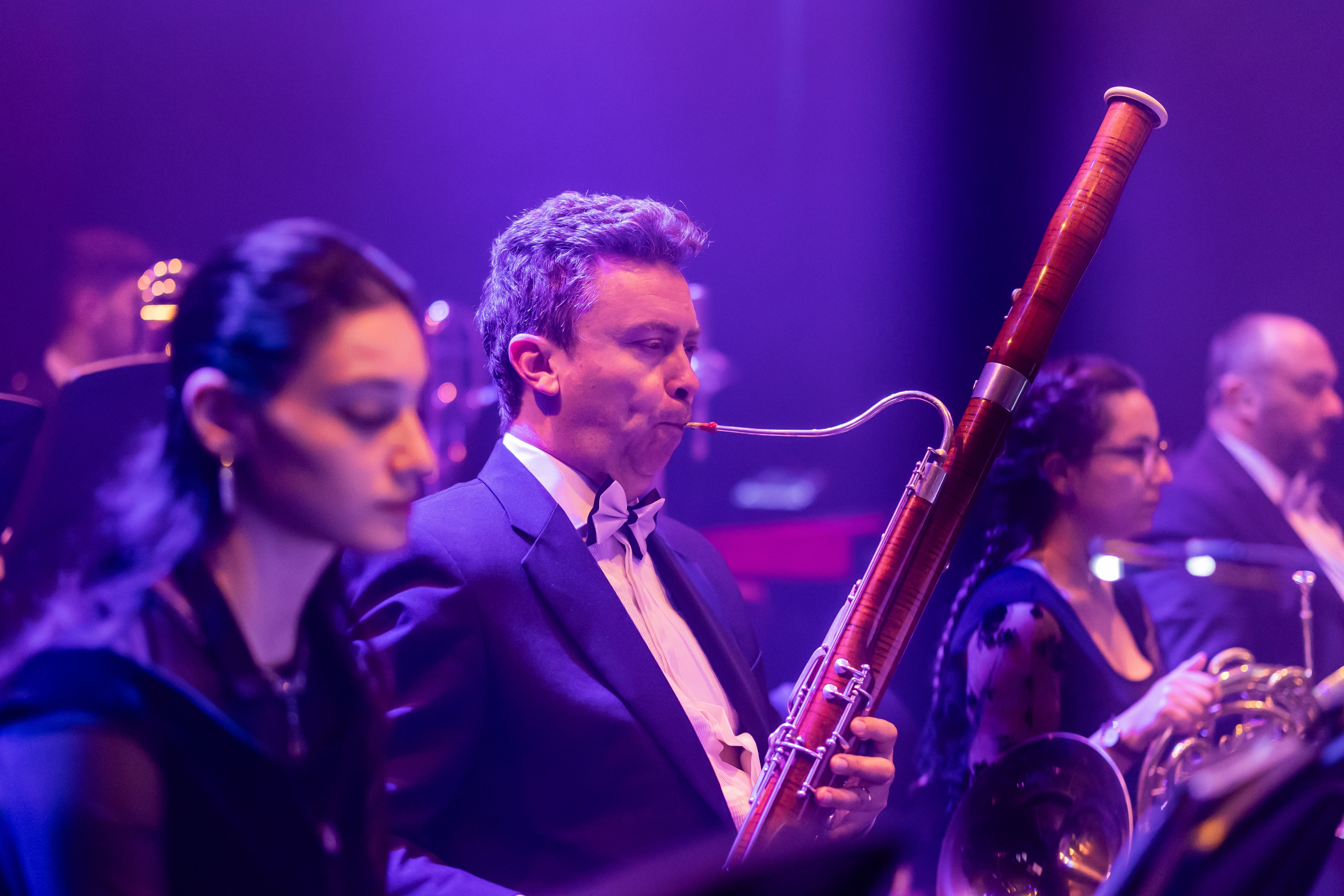 Man in tuxedo playing bassoon with other woodwind and brass musicians next to him.