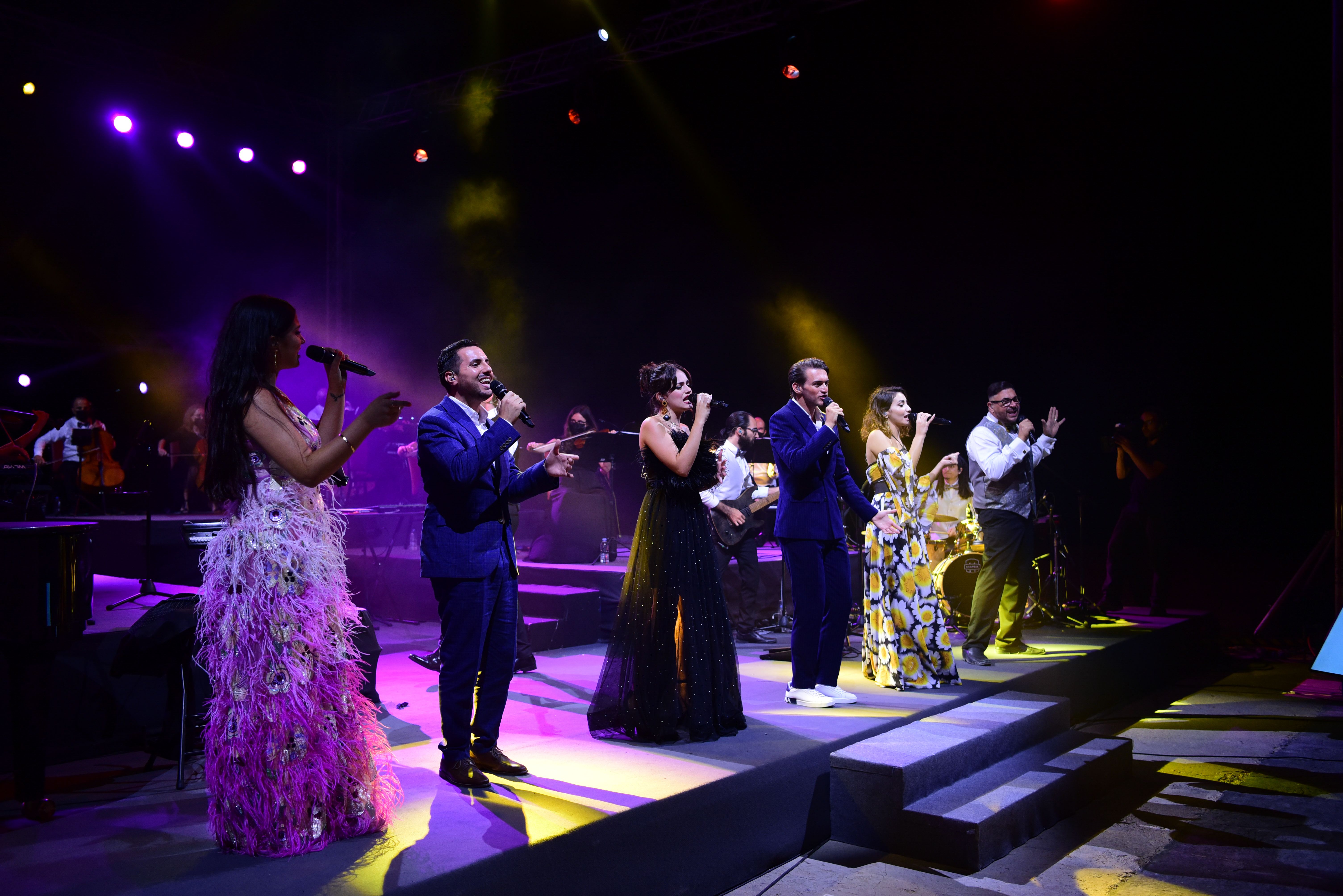 6 popular singers in evening attire holding a mic and singing in a line facing the audience