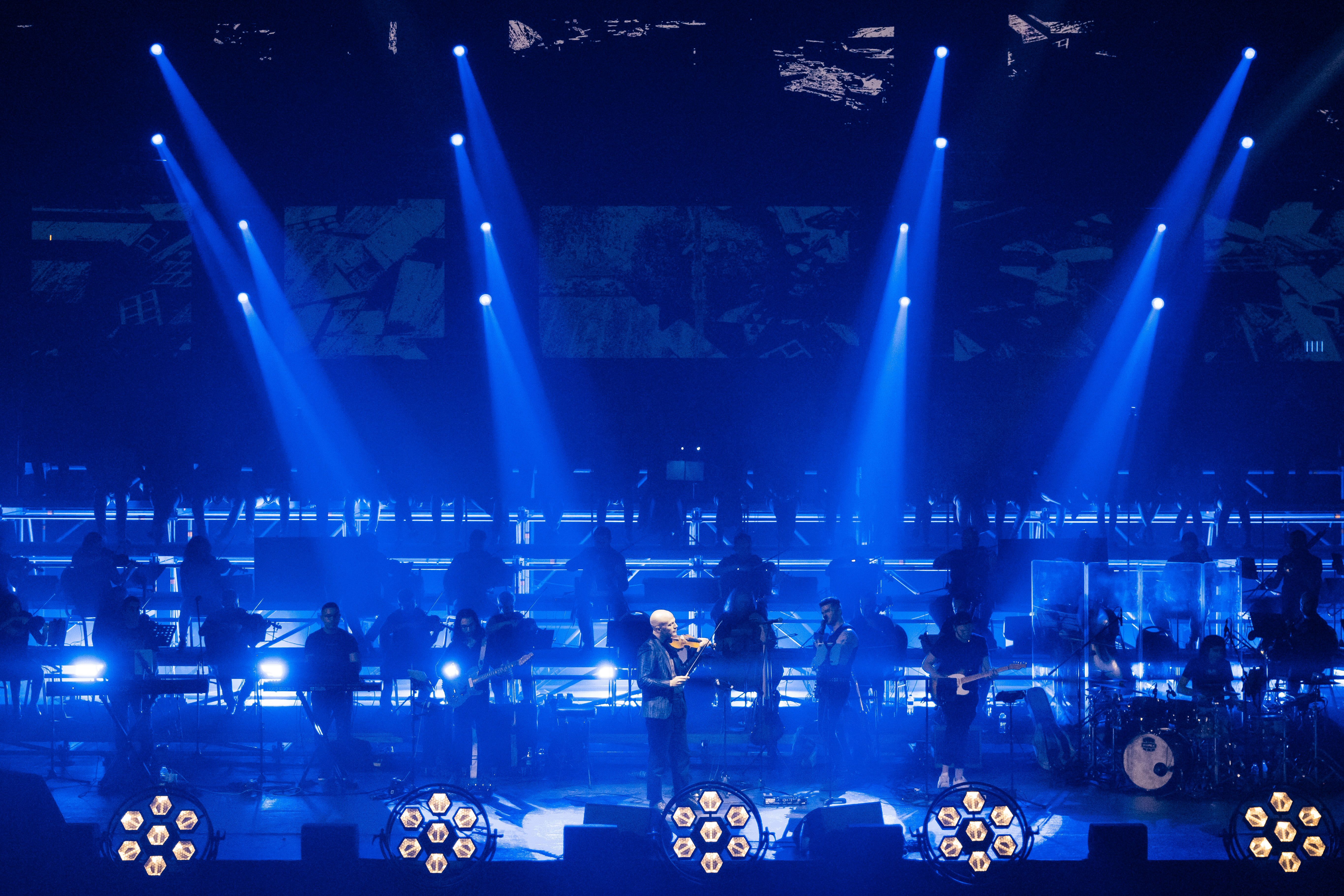 multiple spotlights point down on a stage with a violinist in the foreground and an orchestra behind him