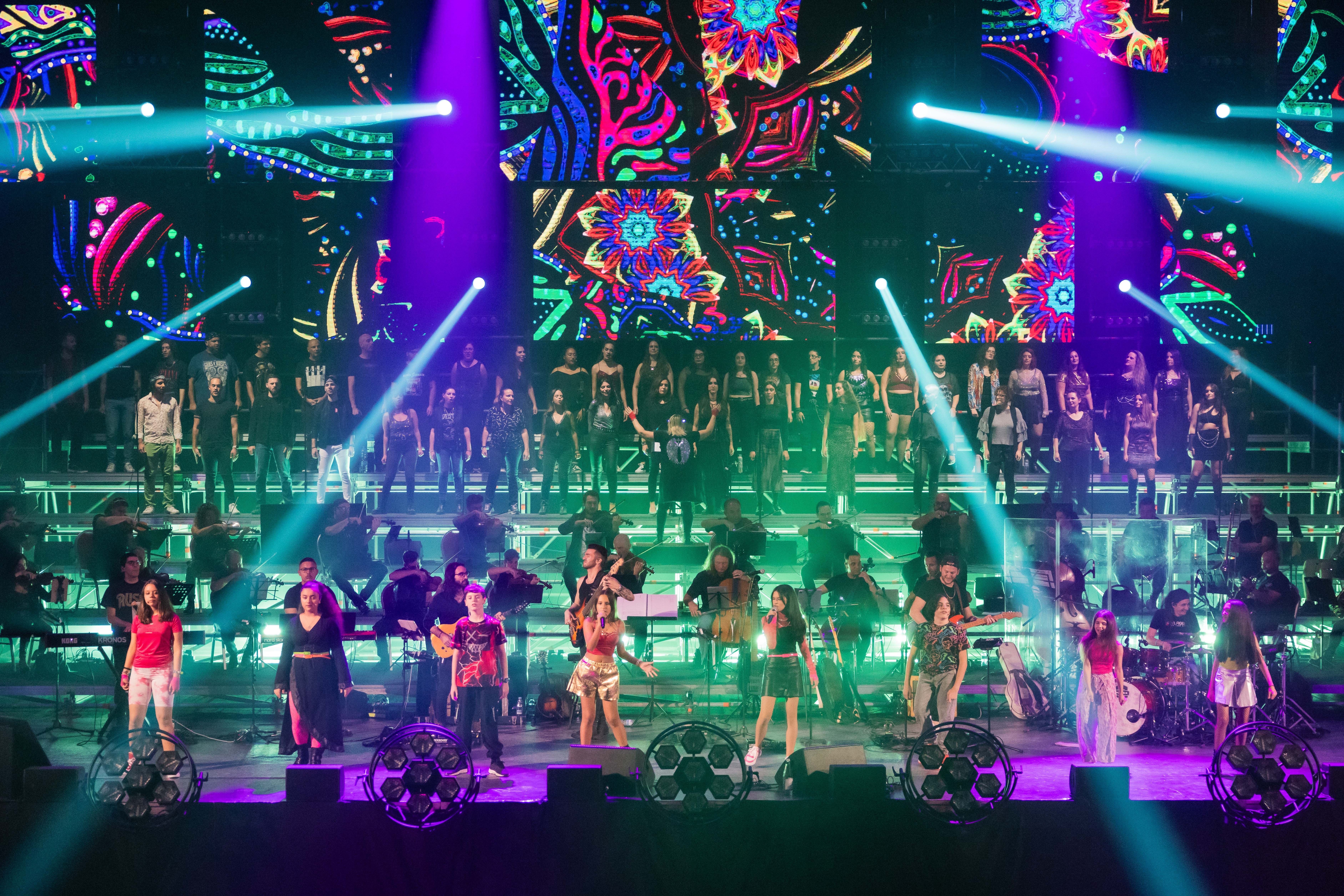 colourful stage with 8 children at the forefront and a band, orchestra and choir in the background