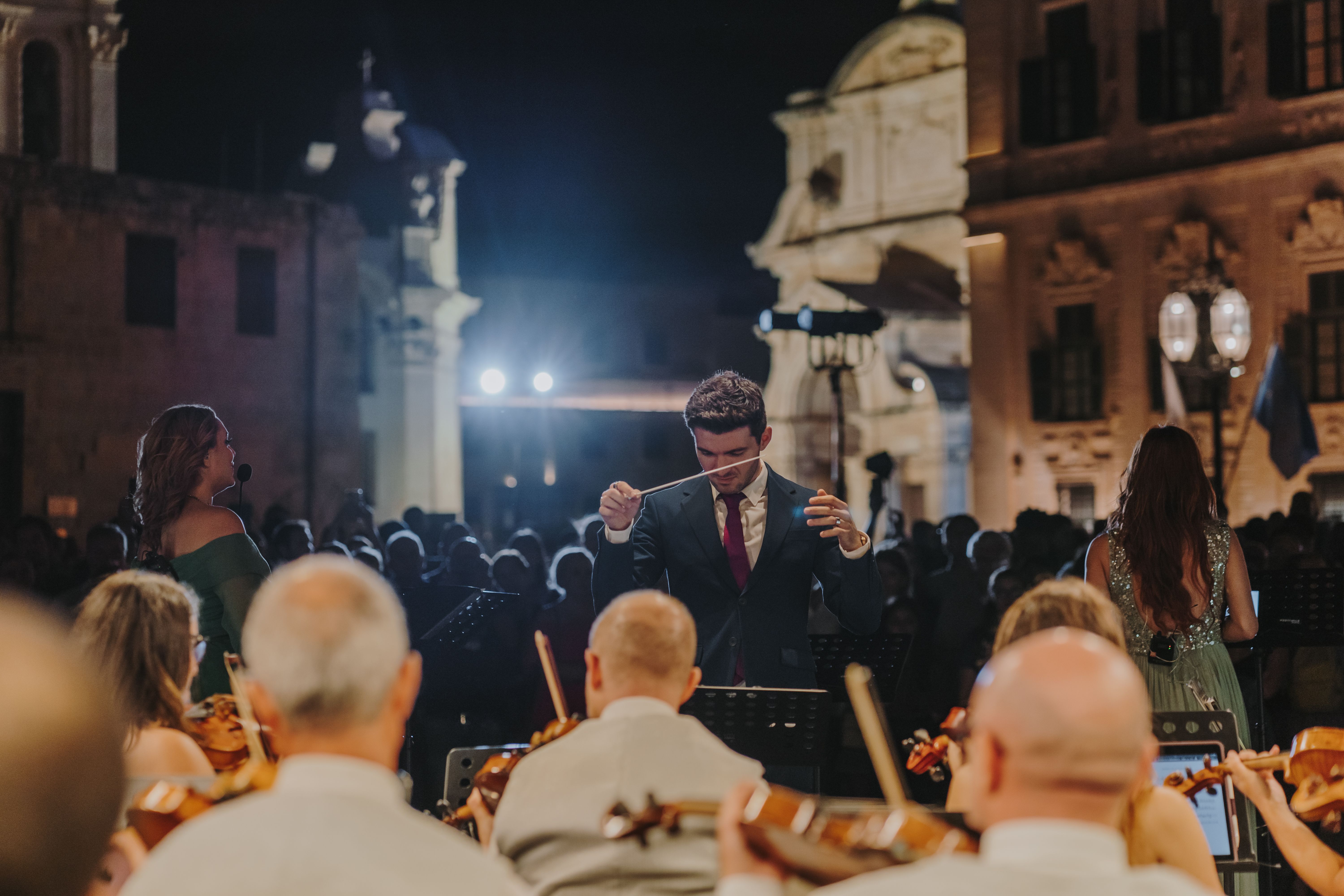 Violinists facing the conductor and singers facing an audience-filled piazza with historical buildings in the background.