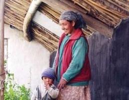 Woman standing with a child beside a vegetable garden outside a traditional Himalayan home.