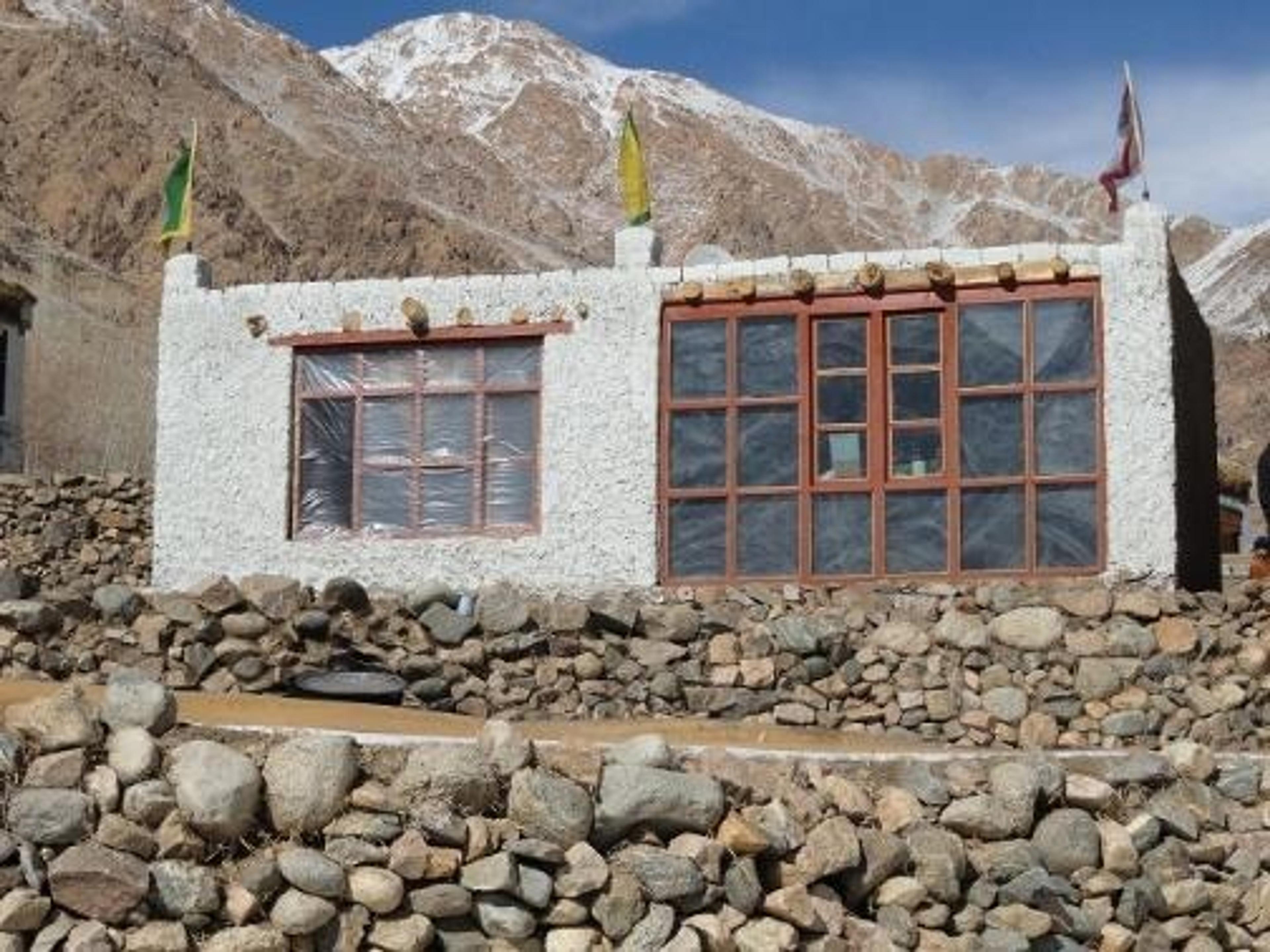 A traditional Himalayan house with large south-facing windows, built among rocky terrain beneath snow-covered mountains.