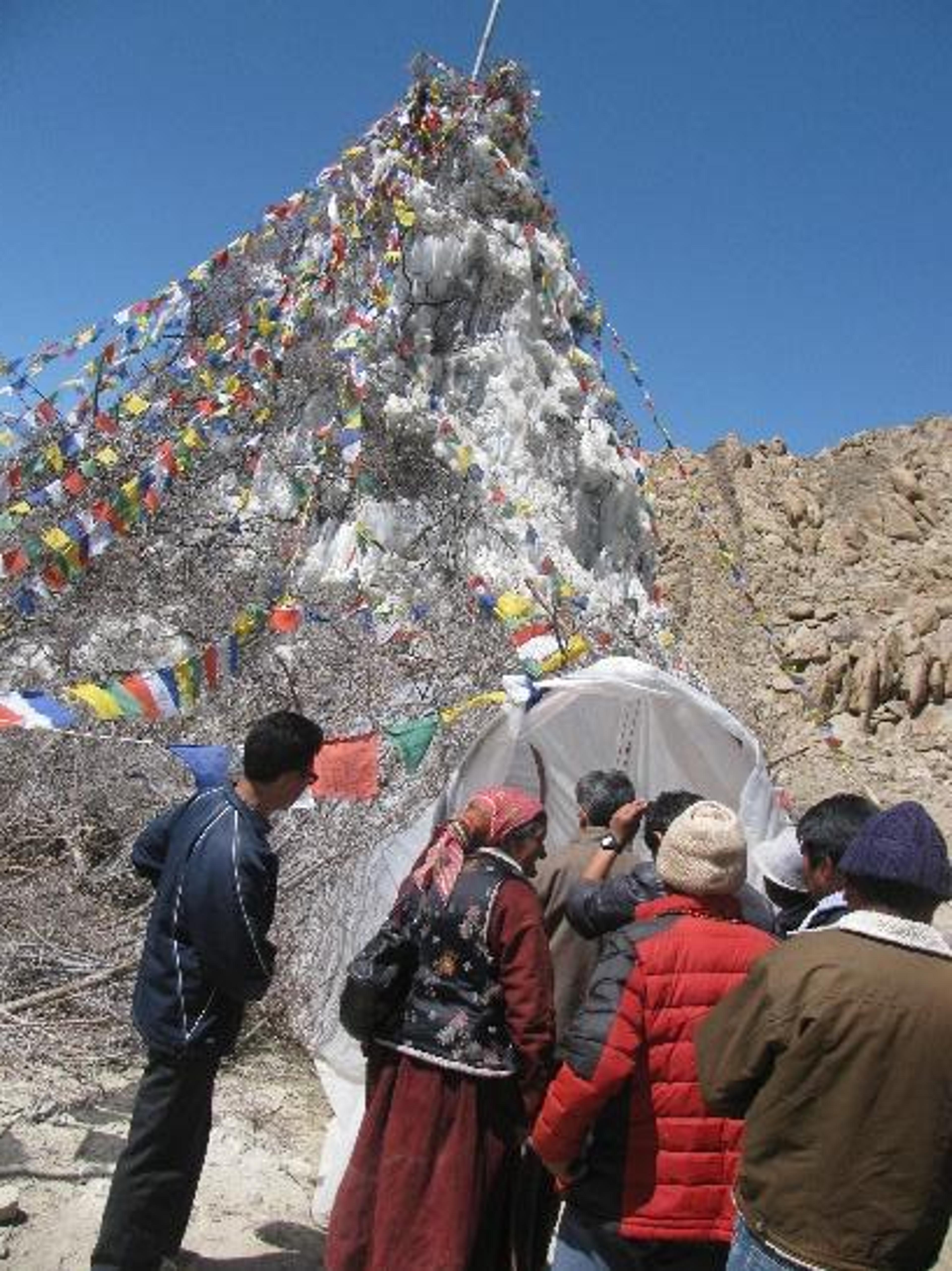 People gathered beside an artificial glacier structure decorated with colorful Tibetan prayer flags in the high-altitude landscape of Ladakh.