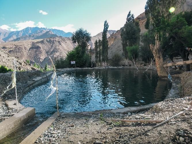 A water storage pond surrounded by stone embankments and tall trees in a mountainous Ladakh landscape, with dry rocky slopes and snow-capped mountains in the background