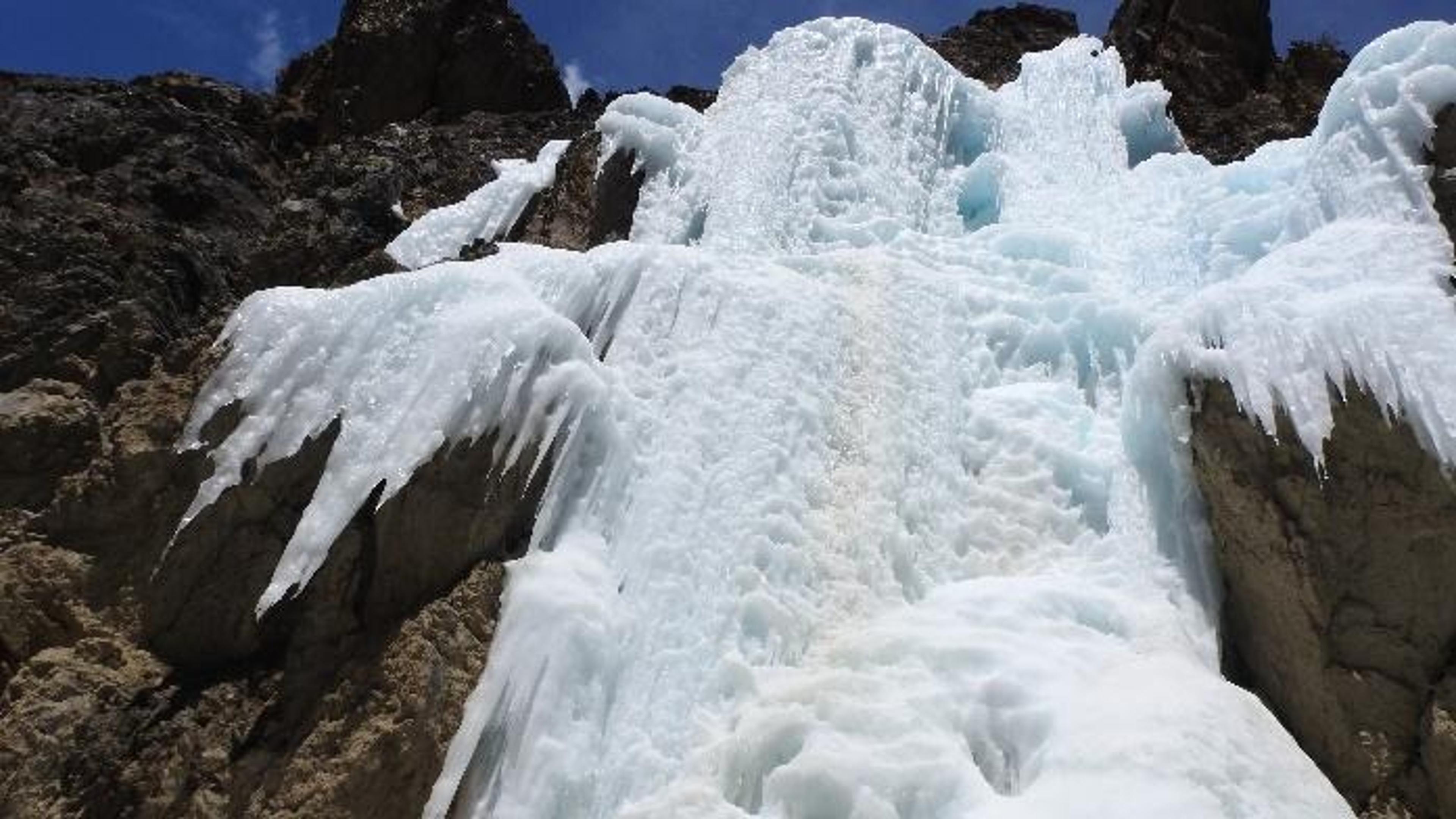 A large formation of blue-white ice covers a rocky mountain slope, with thick frozen layers and icicles extending over dark rocks beneath a clear blue sky.