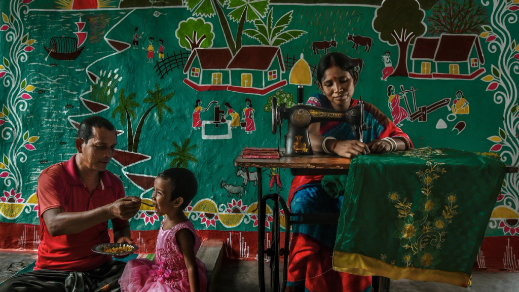 man feeding a small girl while a woman works at a sewing machine in bangladesh