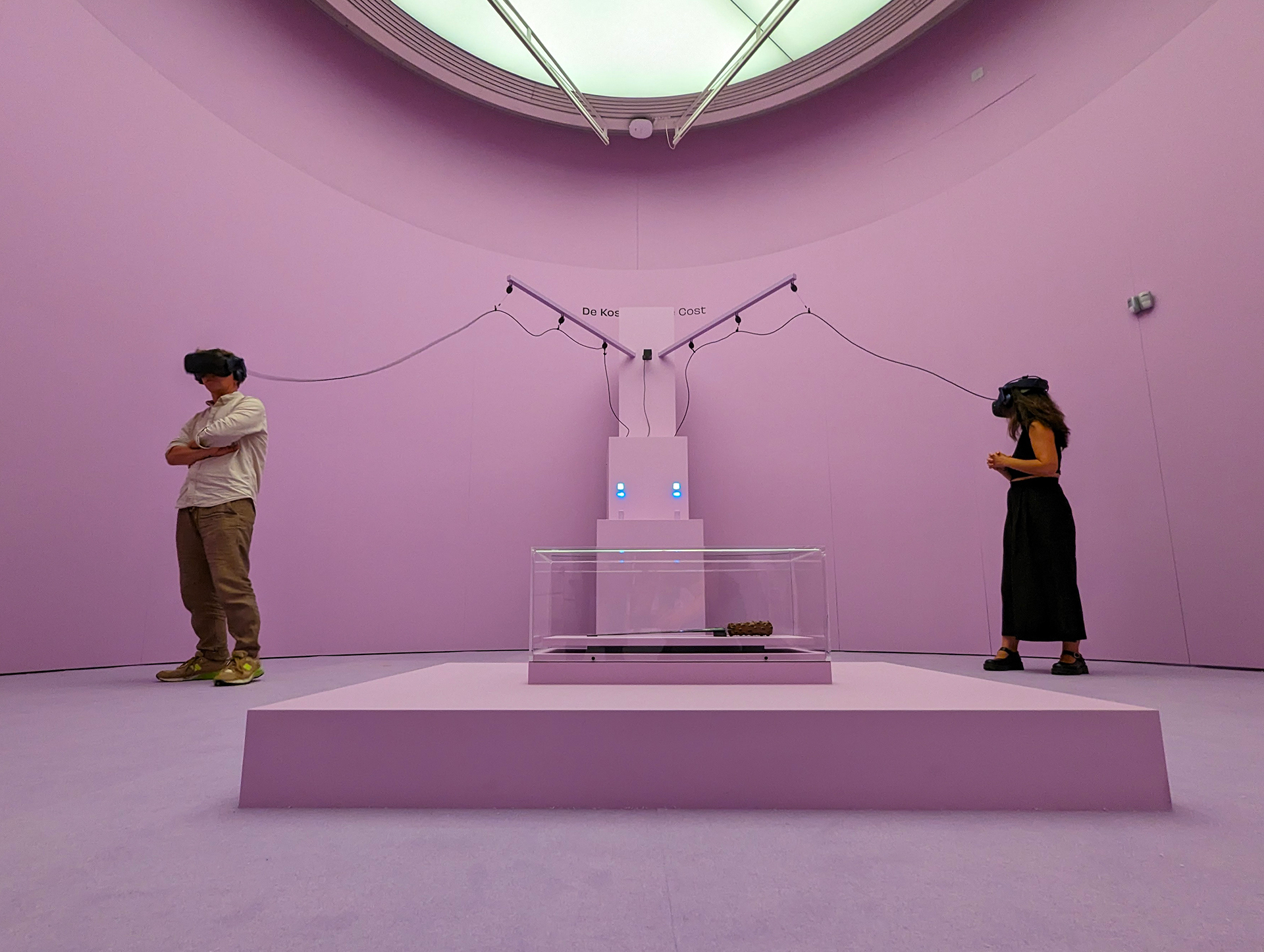 Two visitors wearing VR headsets inside a pink exhibition room around a glass display case