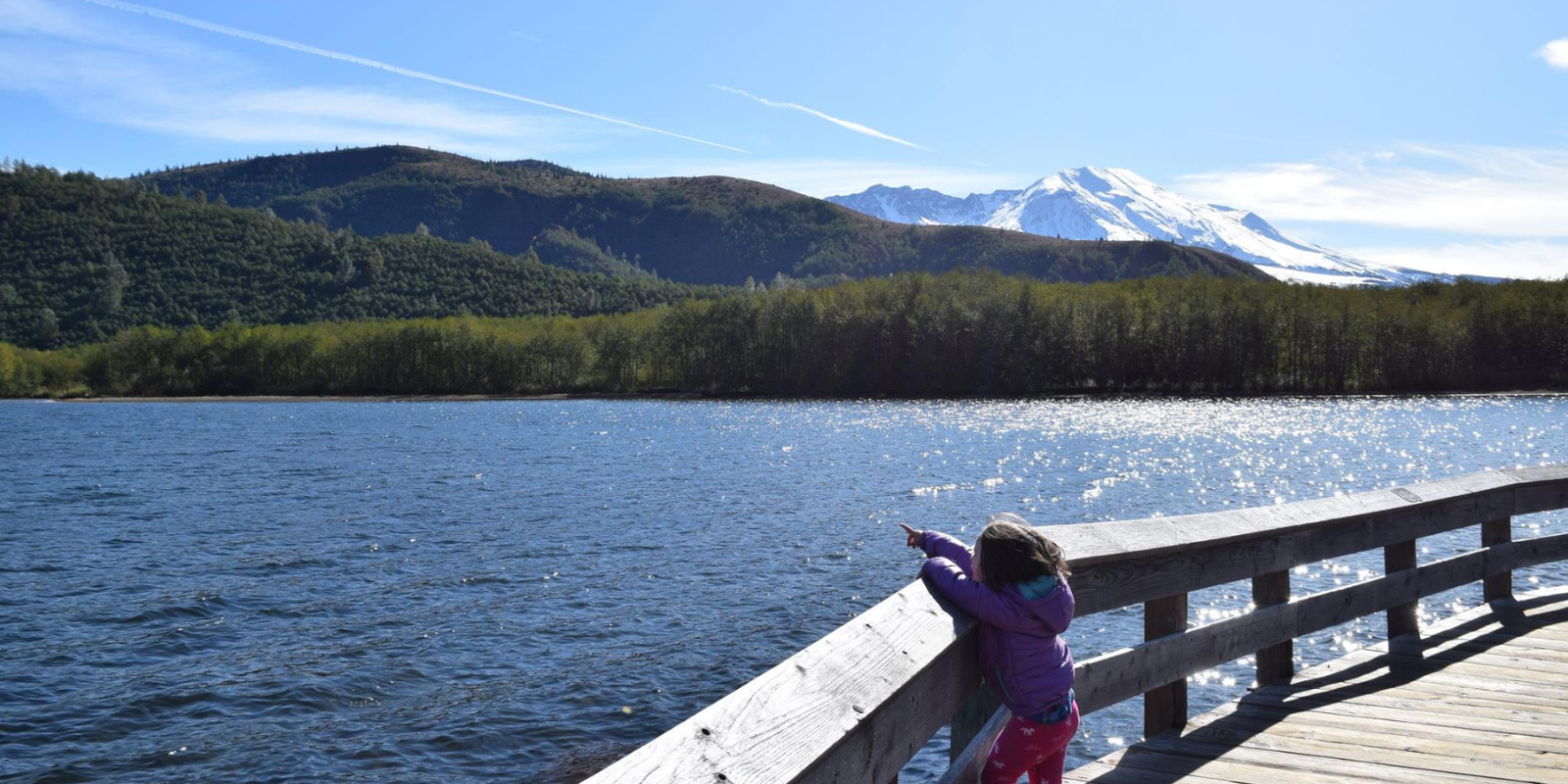 A young child stands on a boardwalk, pointing out at a lake, with Mount St. Helens in the Distance