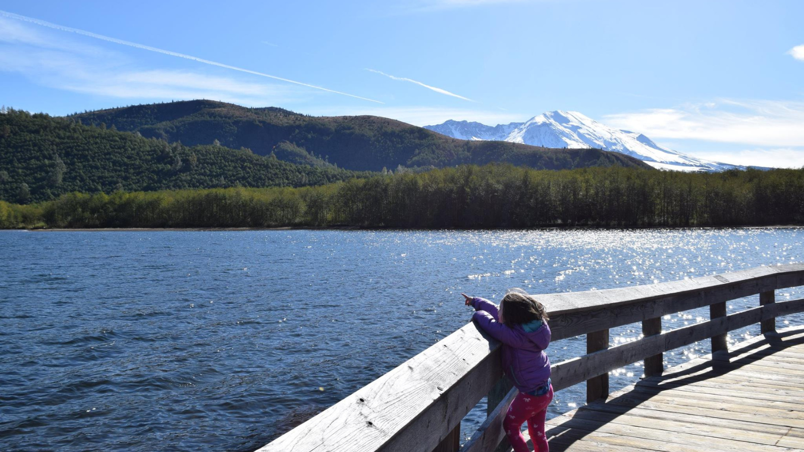 A young child stands on a boardwalk, pointing out at a lake, with Mount St. Helens in the Distance