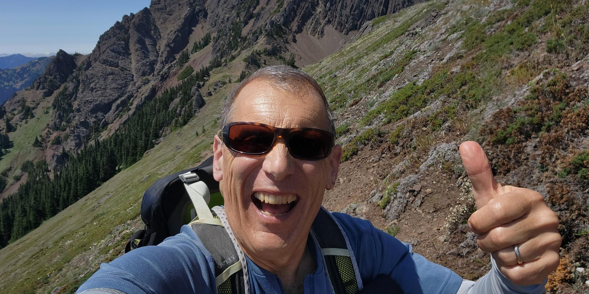 Author and speaker Craig Romano on the trail, giving a thumbs up with mountain peaks int he background.