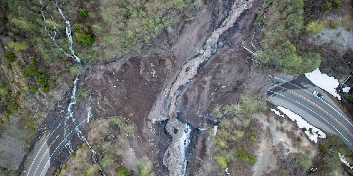 Aerial view of a landslide that has washed out a section of Highway 504 near Mount St. Helens. Mud and debris cover the road, cutting off access across a steep ravine. Forested areas surround the slide, and snow patches are visible near the roadway.