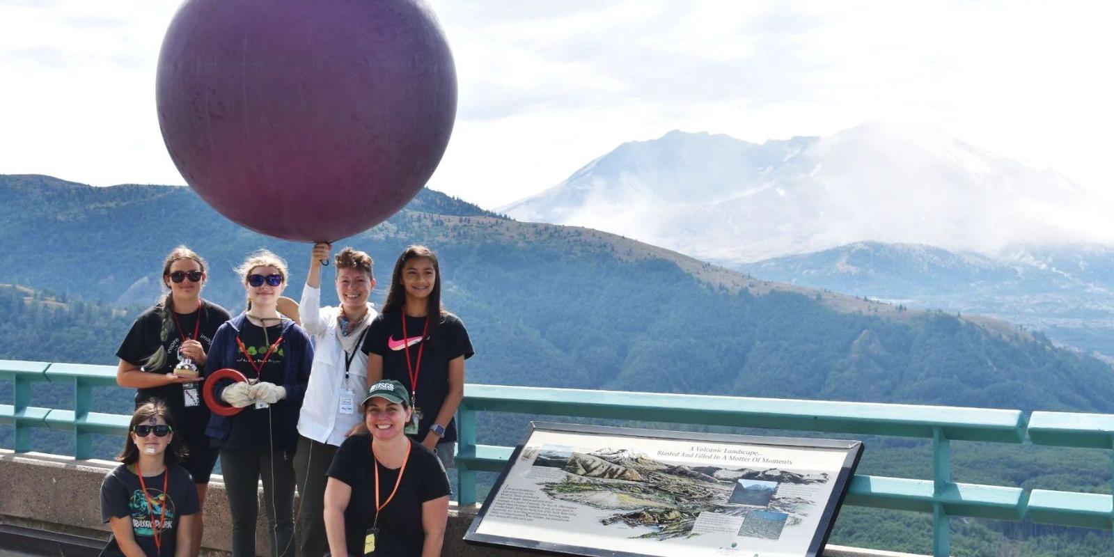 A group of people pose on a scenic overlook with a large weather balloon, preparing for a scientific activity. Behind them, Mount St. Helens is visible in the background, partially shrouded in mist. An interpretive sign in the foreground provides information about the volcanic landscape.