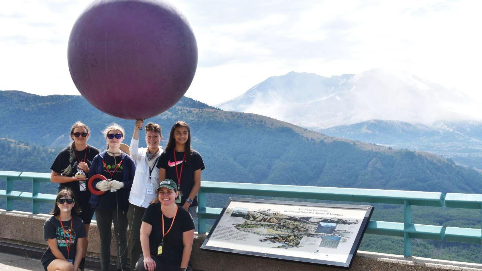 A group of people pose on a scenic overlook with a large weather balloon, preparing for a scientific activity. Behind them, Mount St. Helens is visible in the background, partially shrouded in mist. An interpretive sign in the foreground provides information about the volcanic landscape.
