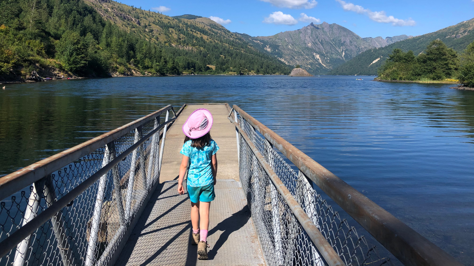 A young child walks on a boardwalk toward a lake with forested rolling hills in the background