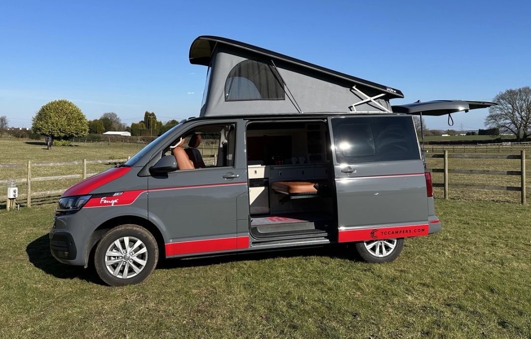 Fenyx, a grey VW Transporter campervan with red detailing, the pop-top roof up and the side door open to the interior, parked on grass