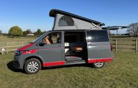 Fenyx, a grey VW Transporter campervan with red detailing, the pop-top roof up and the side door open to the interior, parked on grass