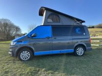 Enola, a grey VW Transporter campervan with blue detailing and the pop-top roof raised, parked on grass