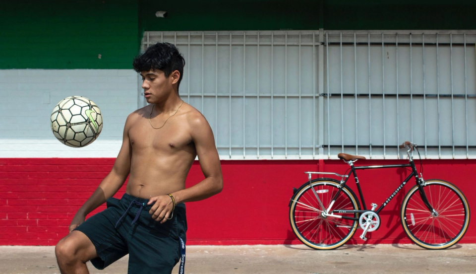 A young footballer controls a football in mid-air while practicing outdoors in daylight. He stands shirtless in front of a colorful building with green, white, and red walls, with a bicycle parked behind him.