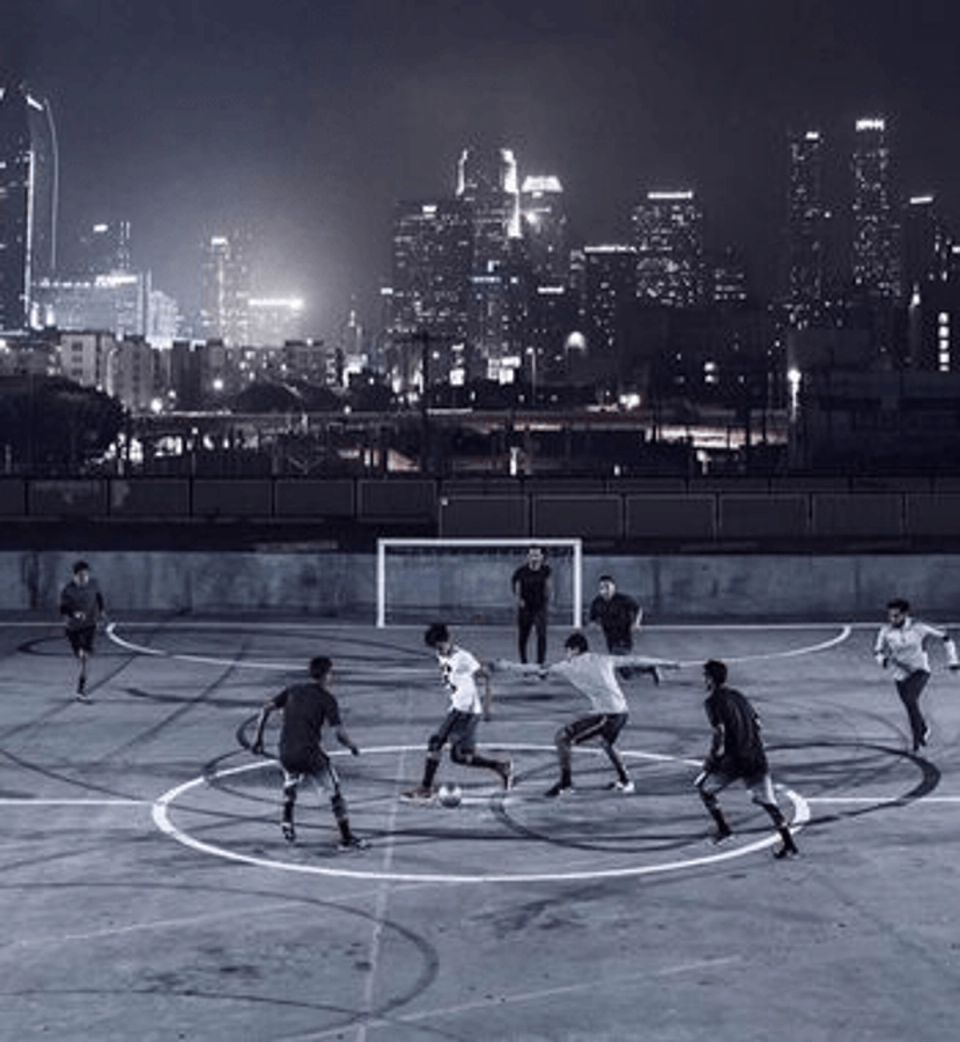 Footballers play a fast-paced small-sided match on an outdoor hard court at night, with a brightly lit city skyline in the background, illustrating the dynamic and unpredictable nature of the game.