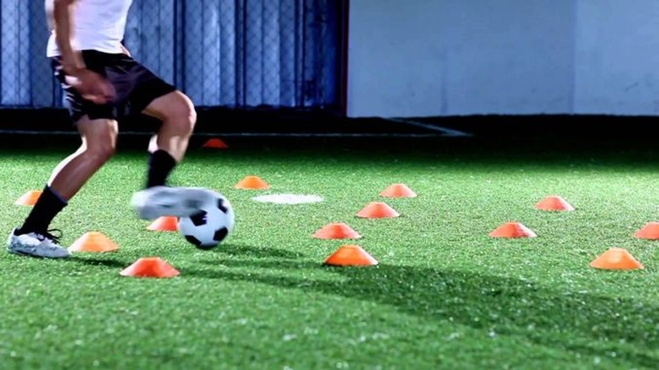 A footballer dribbles a ball through a line of orange cones during an isolated training drill on artificial turf under bright lights.