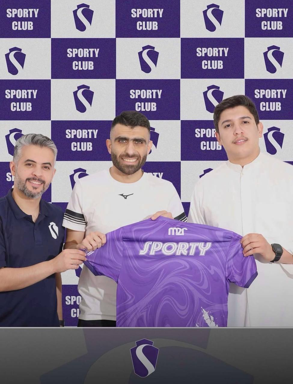 Professional footballer Ibrahim Tamimi poses with two Sporty Club representatives while holding a purple club jersey after signing with his new team, standing in front of the club's branded backdrop.