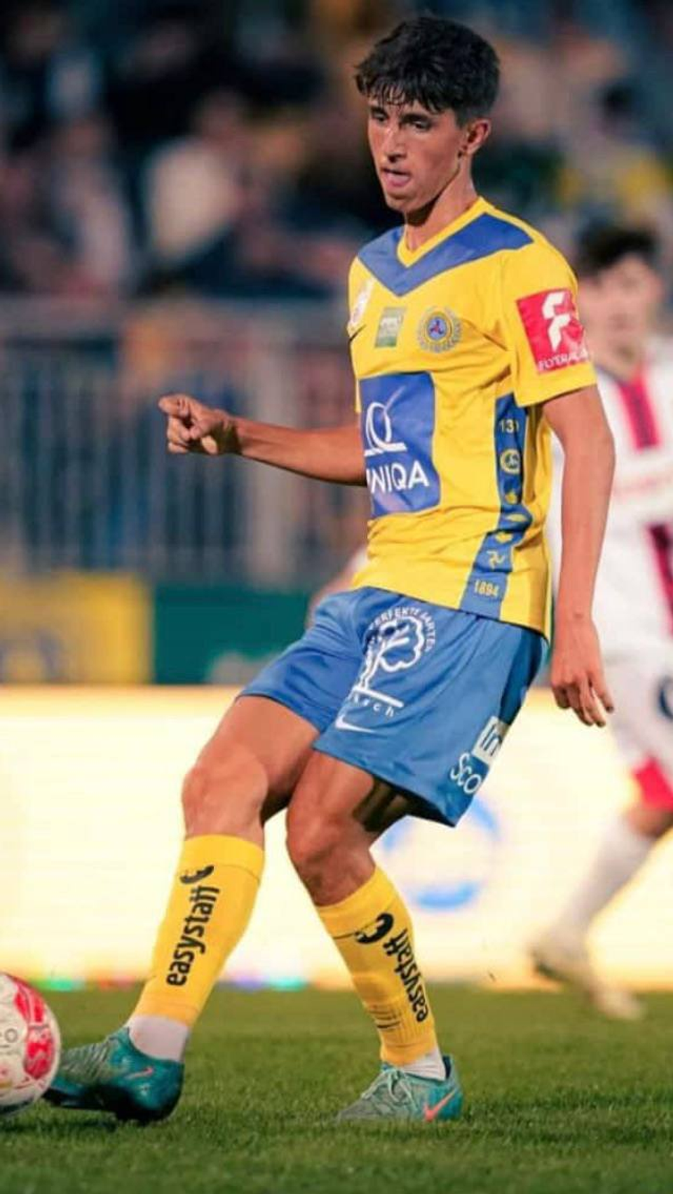 Professional footballer Dean Titkov of First Vienna Football Club 1894 during a match, controlling the ball in a yellow and blue kit.