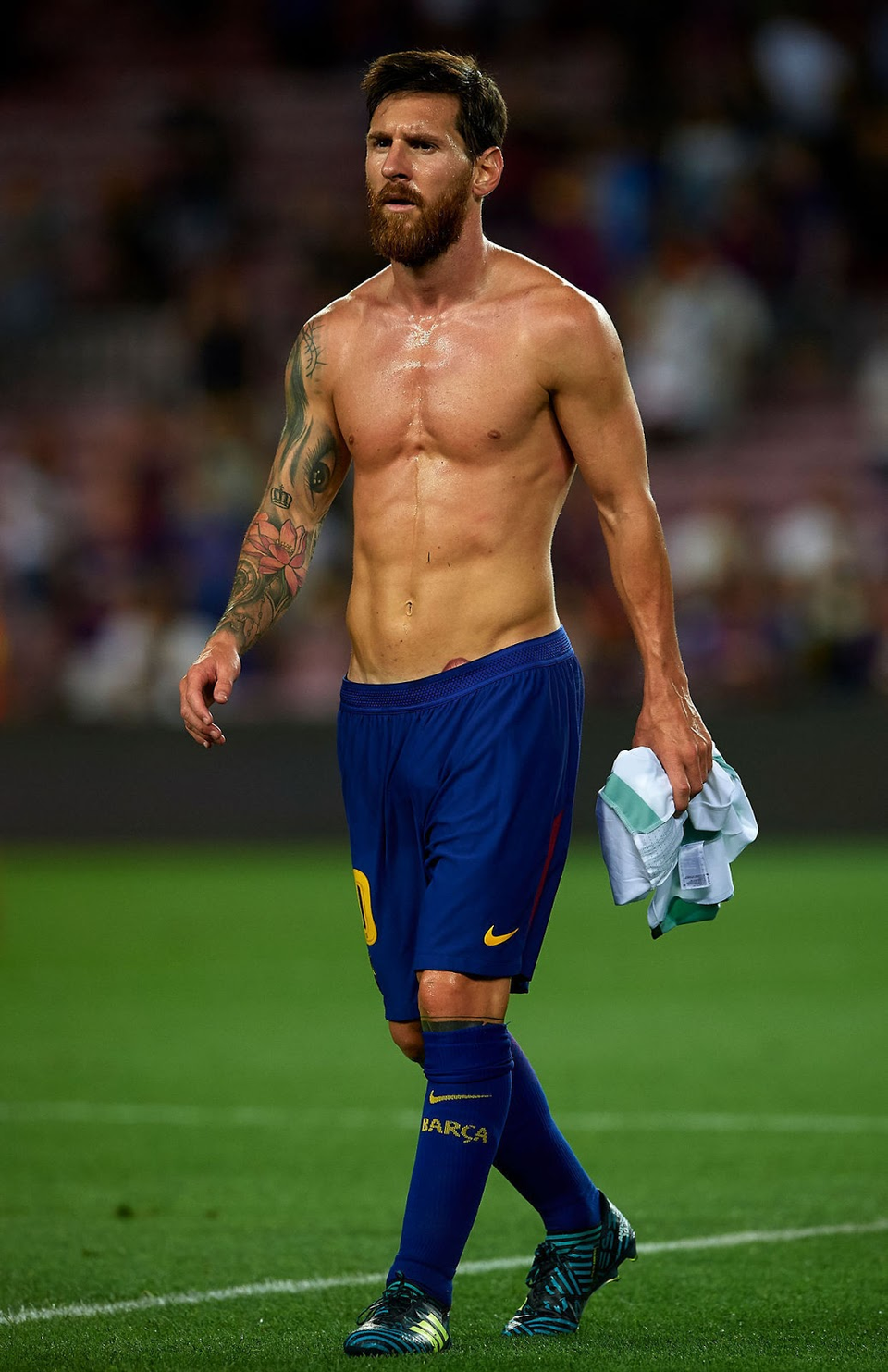 Lionel Messi walks across the pitch shirtless after a match, displaying a lean, athletic physique developed for elite football performance, with Barcelona shorts and socks still on.