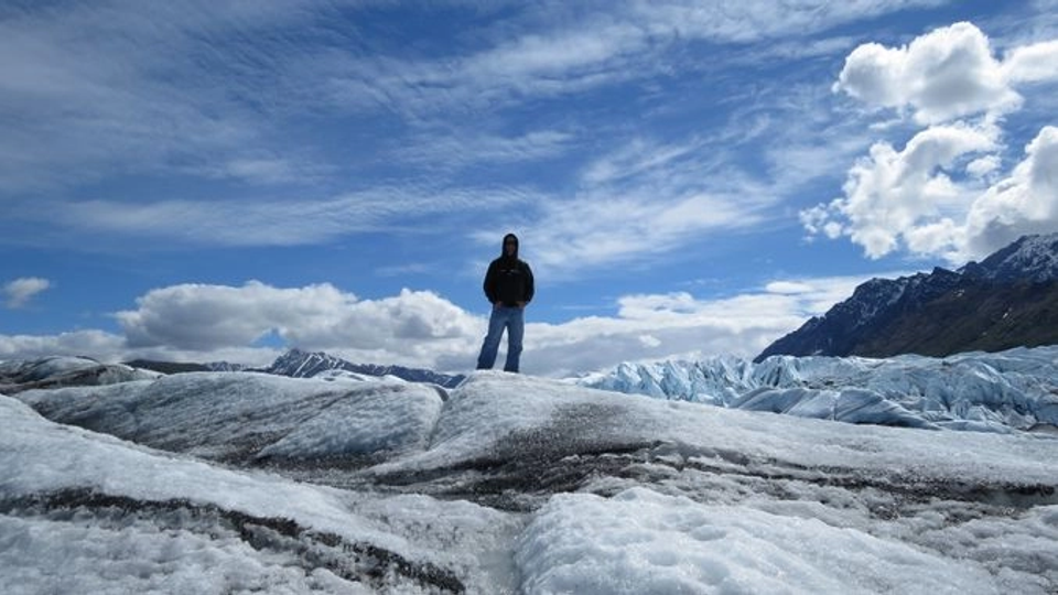 Person standing alone on a glacier beneath a bright blue sky with scattered clouds, surrounded by snow-covered mountains and expansive icy terrain.